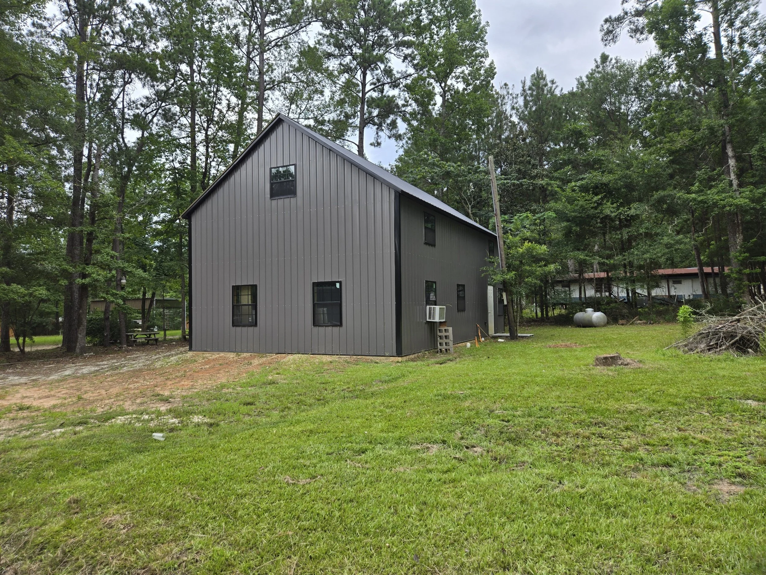 A gray two-story building with a steep gable roof, black-framed windows, an air conditioning unit on the side, set on a grassy yard with some trees and propane tanks in the background.