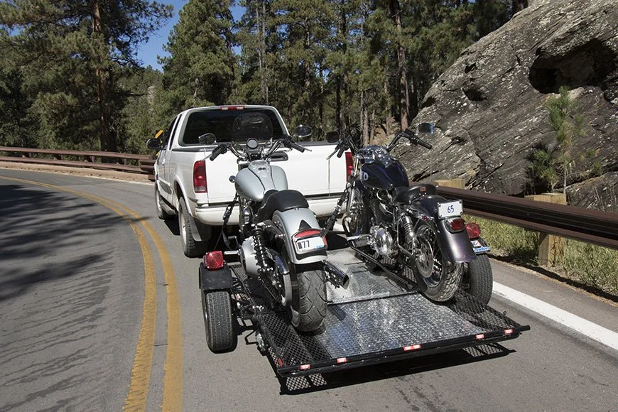 A white pickup truck towing two motorcycles on a flatbed trailer on a mountain road with pine trees and rocks on either side.