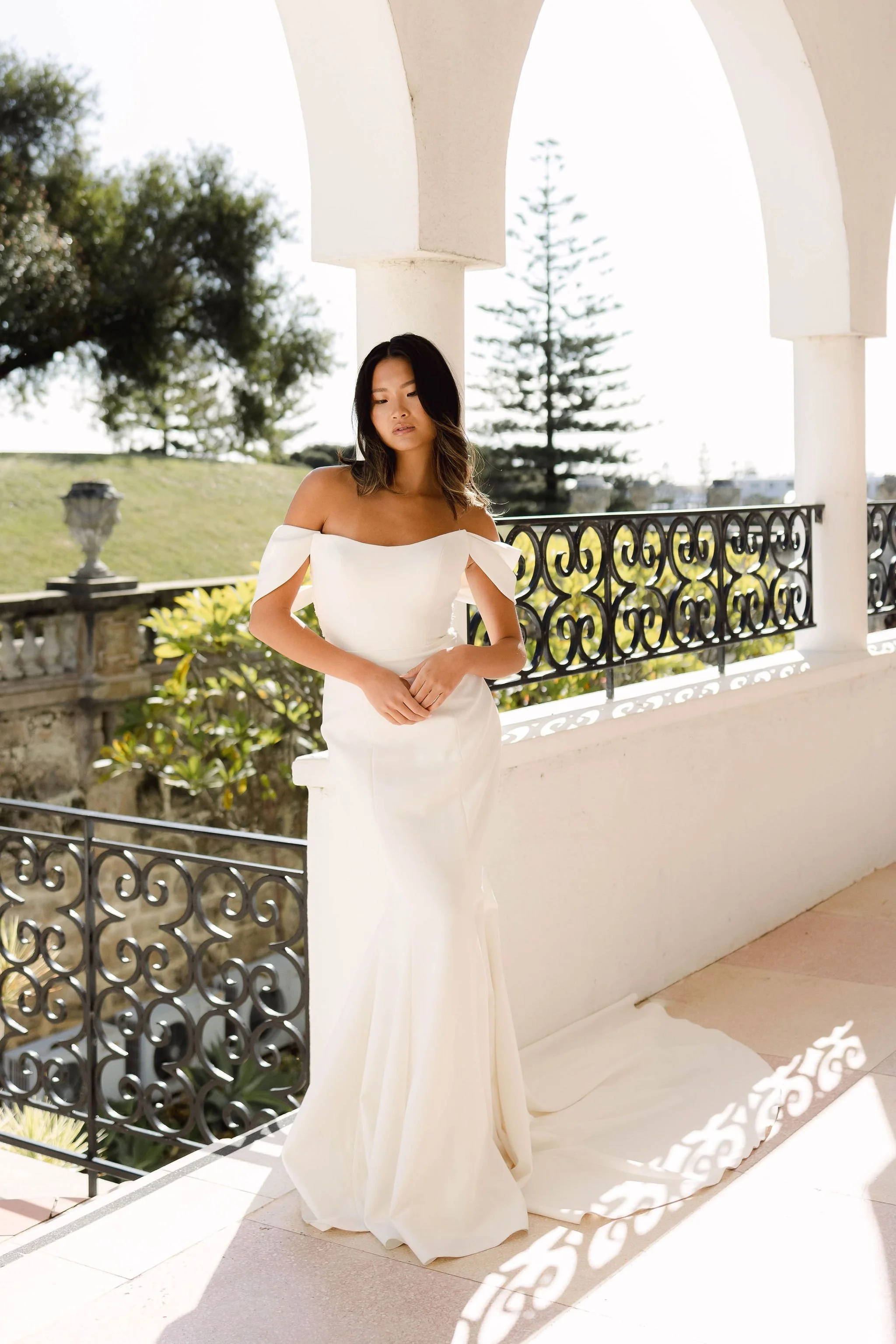 Woman in an off-shoulder white wedding dress standing on a balcony with ornate metal railings, surrounded by greenery and architectural details.