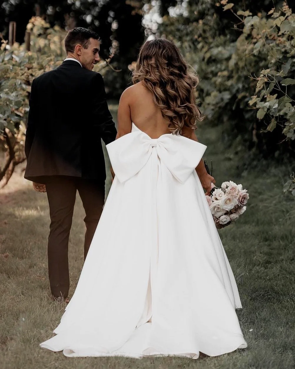Bride and groom walking outdoors, bride in a white dress with a large bow, holding a bouquet.