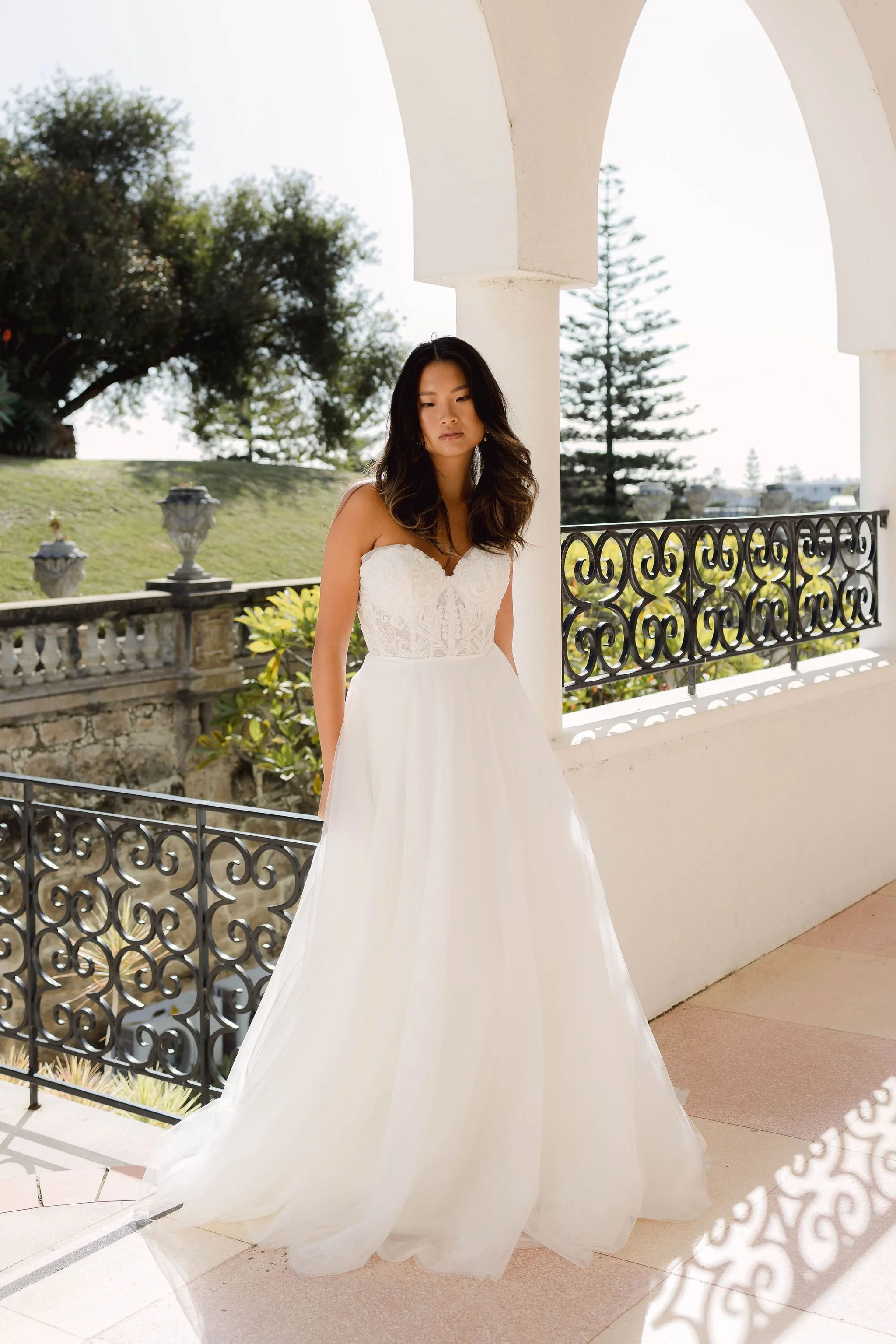 Woman in a strapless white wedding dress standing on a balcony with decorative iron railing, outdoor setting with trees in the background.