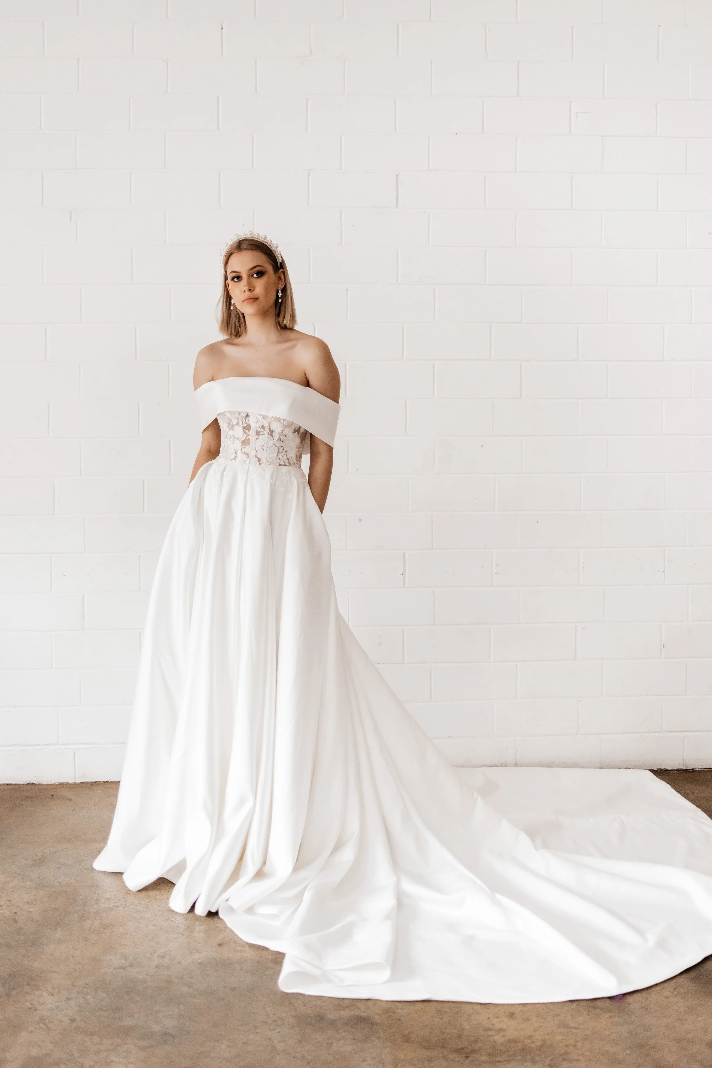 Bride in an off-shoulder white wedding dress with lace bodice, standing against white brick wall.