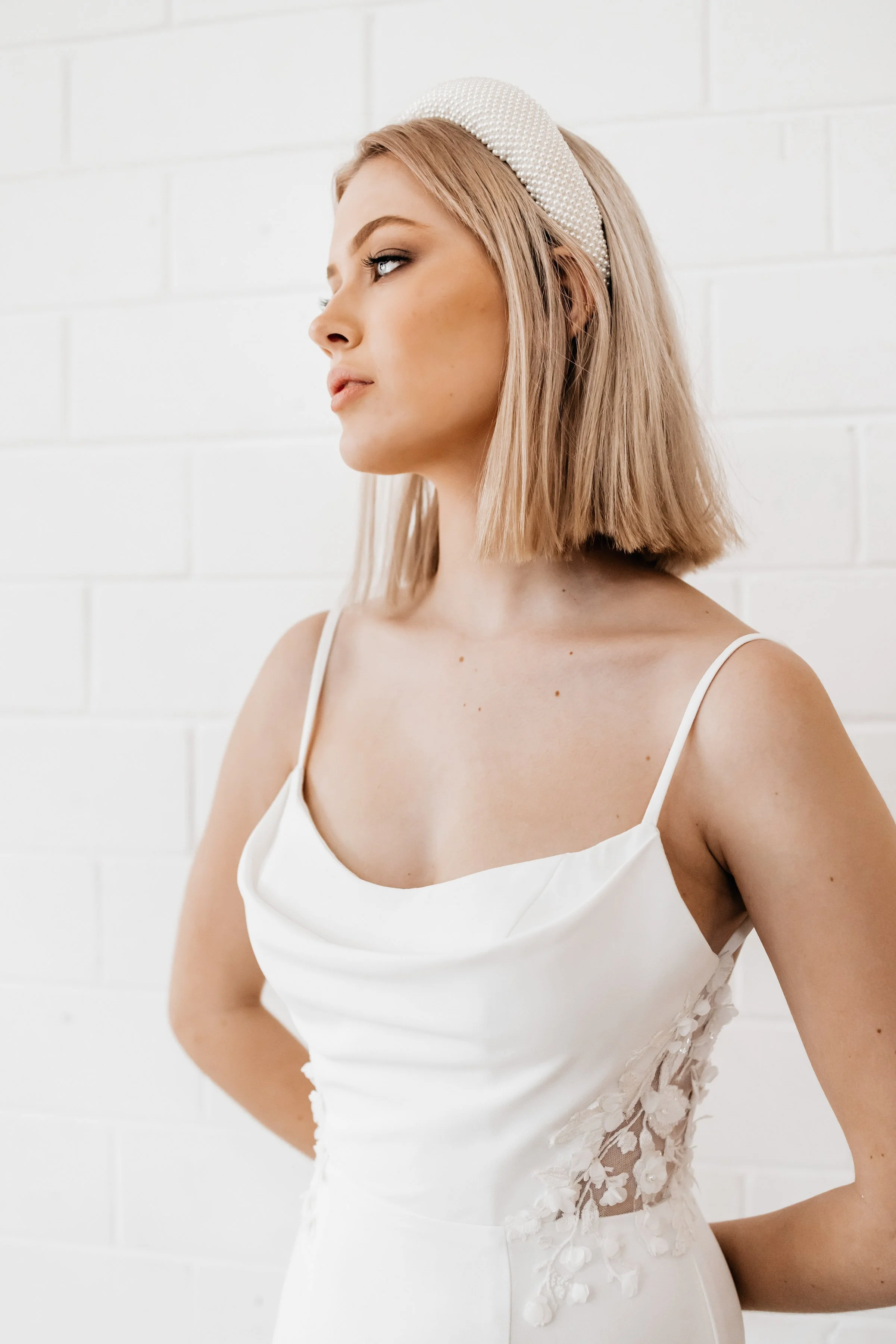 Woman in a white sleeveless dress with floral lace details, wearing a pearl headband, standing against a white brick wall.