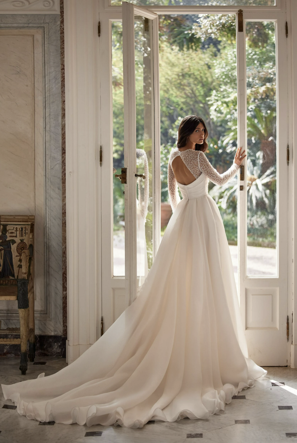 Bride in elegant white wedding gown with lace long sleeves, standing by open French doors, looking outside.