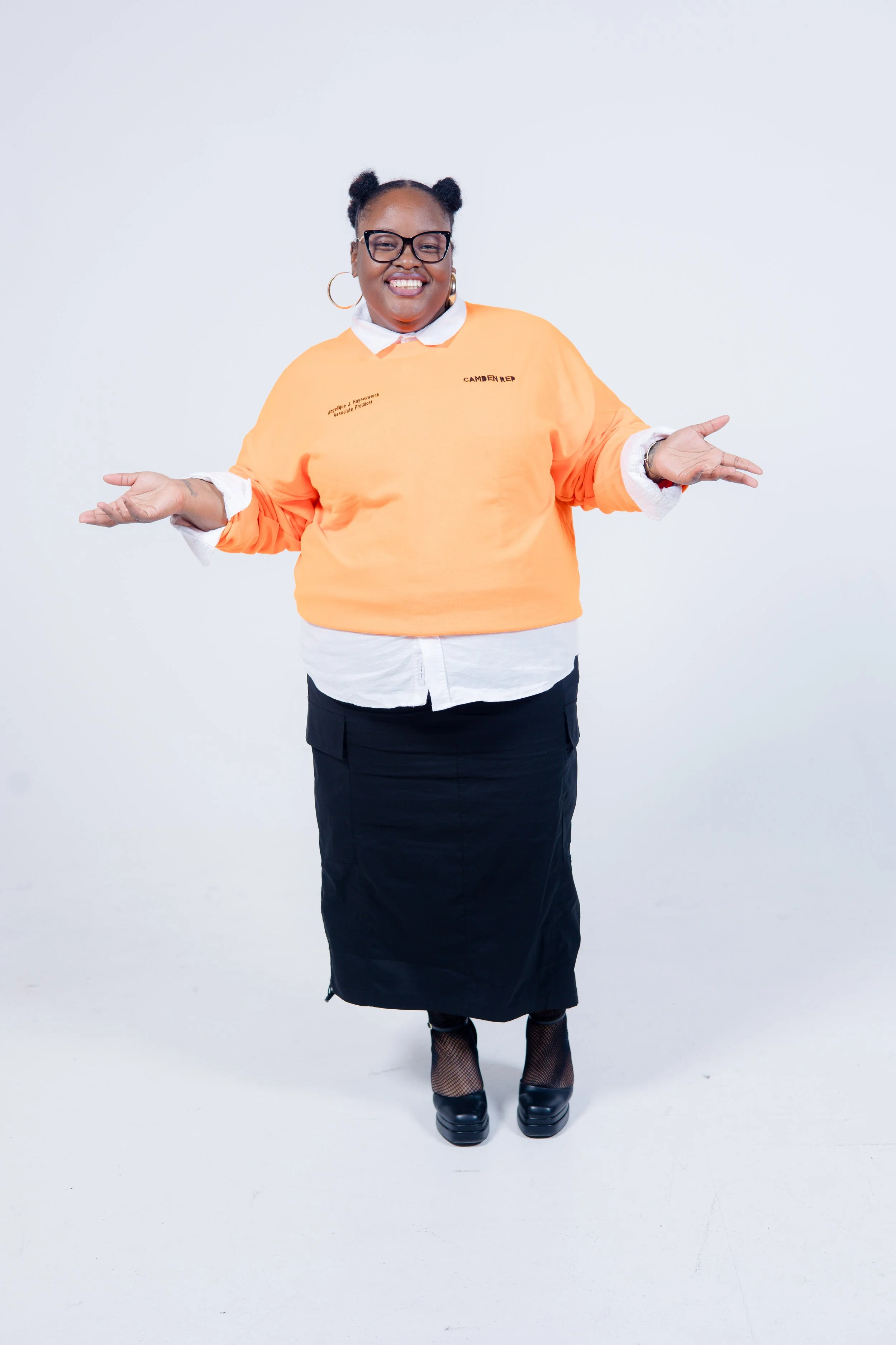 Person standing in a studio, wearing an orange sweater, black skirt, and glasses, smiling with arms open.