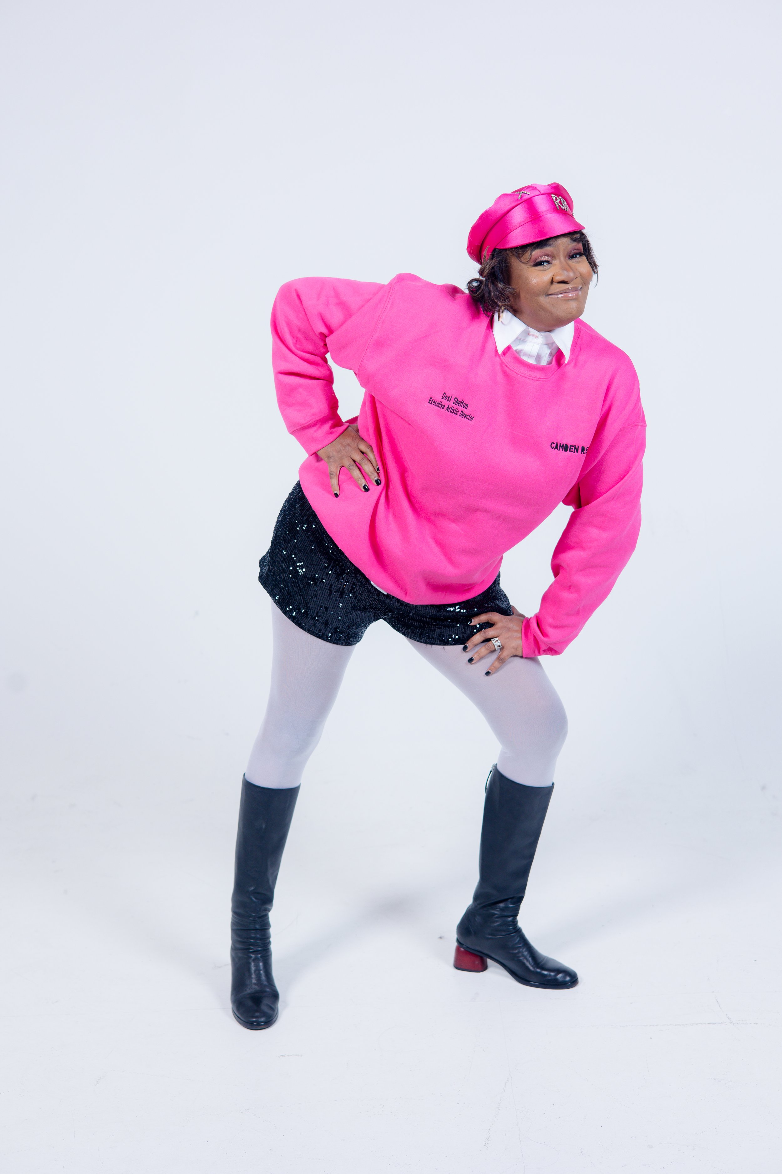 Person wearing a pink sweatshirt, black sequined shorts, white tights, black boots, and a pink beret, posing against a plain background.