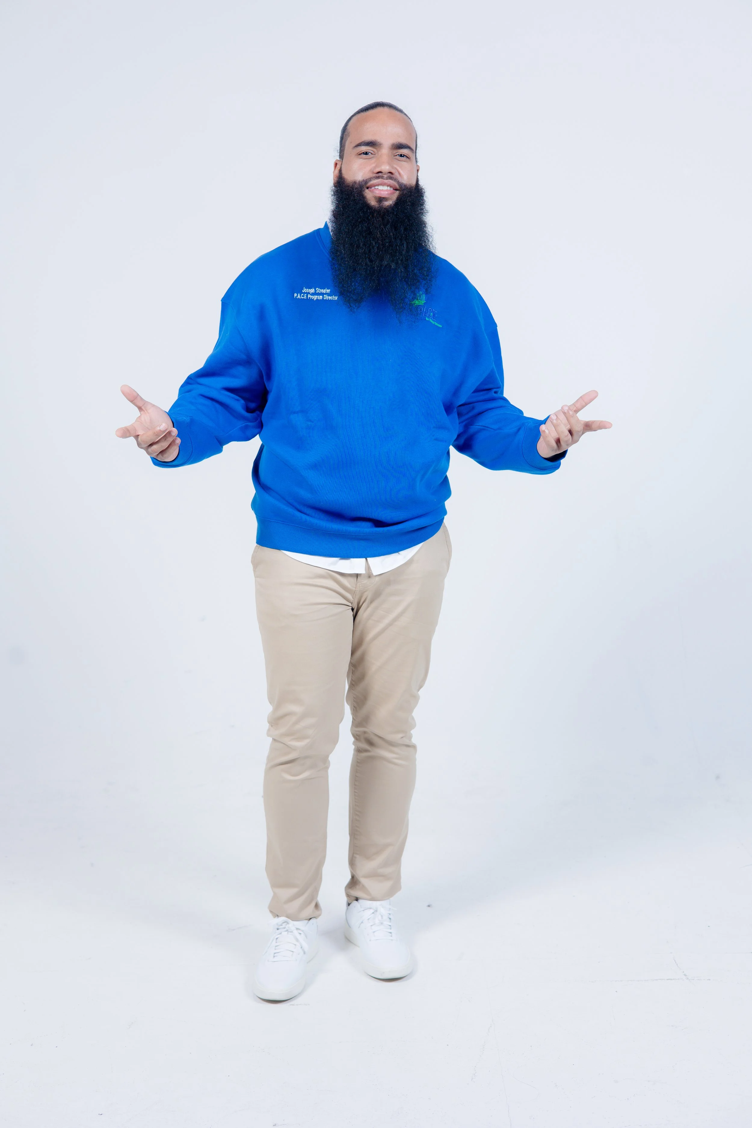 A man with a long beard wearing a blue sweater, beige pants, and white sneakers, standing with arms open on a white background.