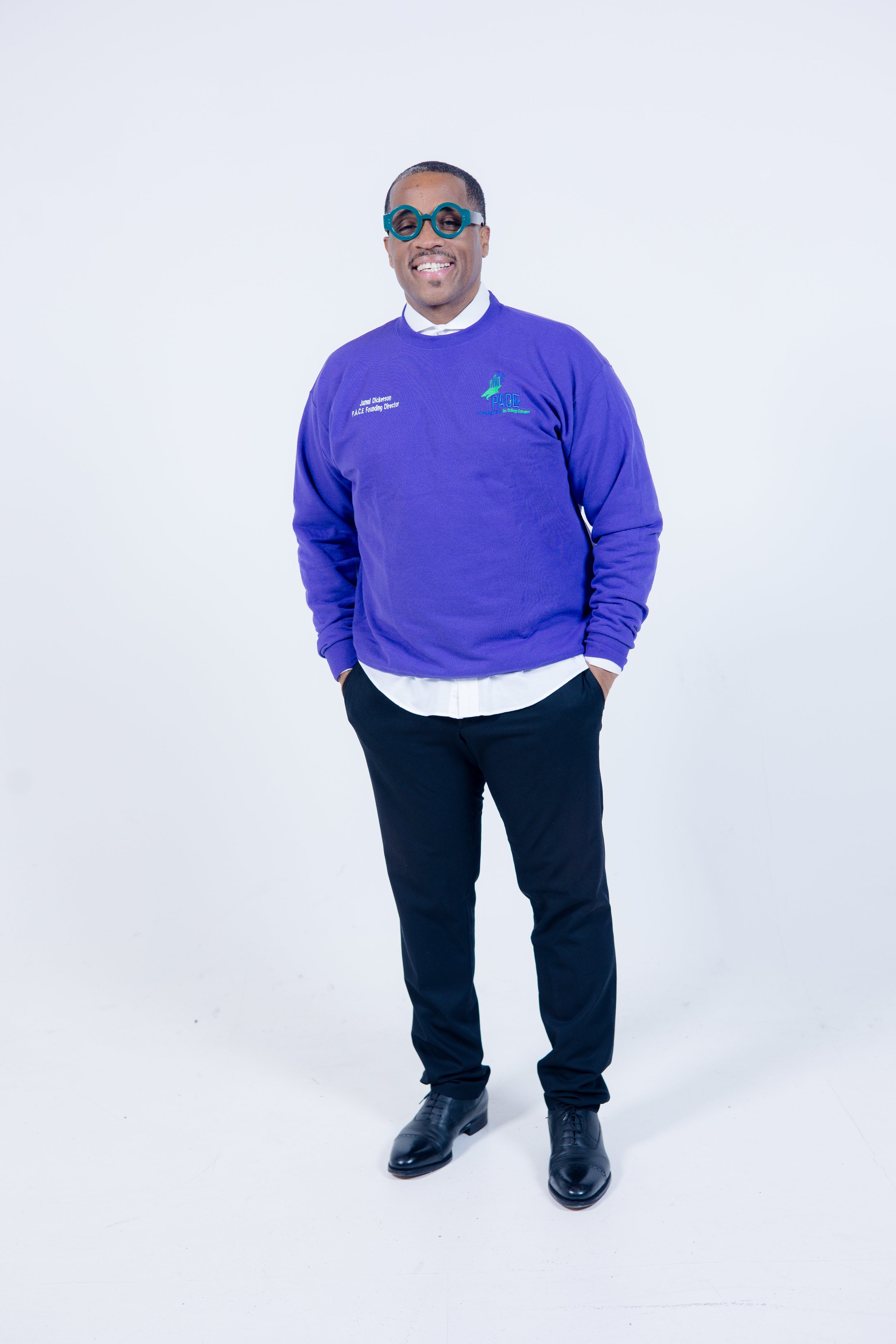 Smiling man in a purple sweatshirt with text, black pants, and black shoes standing against a white background.