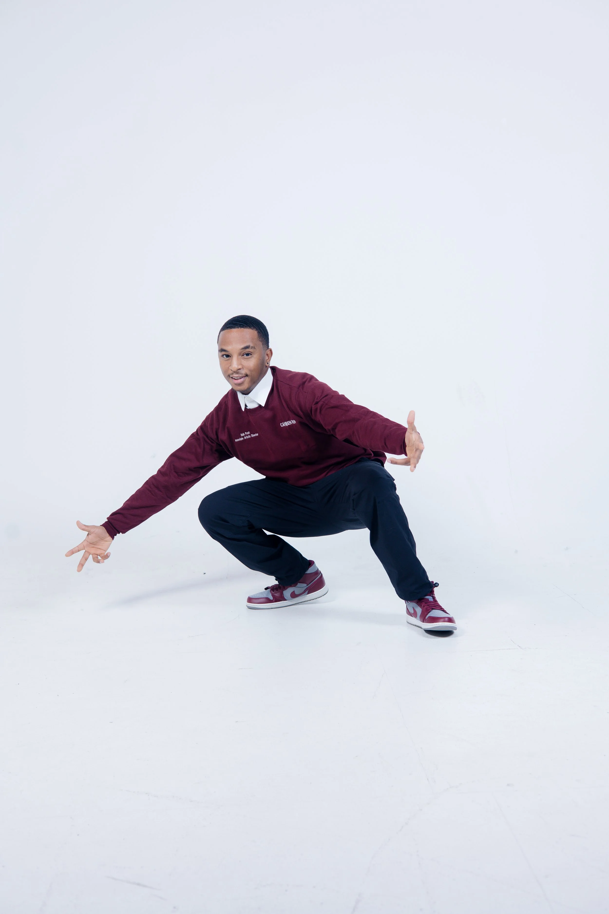 A person squatting with arms extended outward, wearing a maroon sweater, white shirt, black pants, and maroon and white sneakers against a light background.