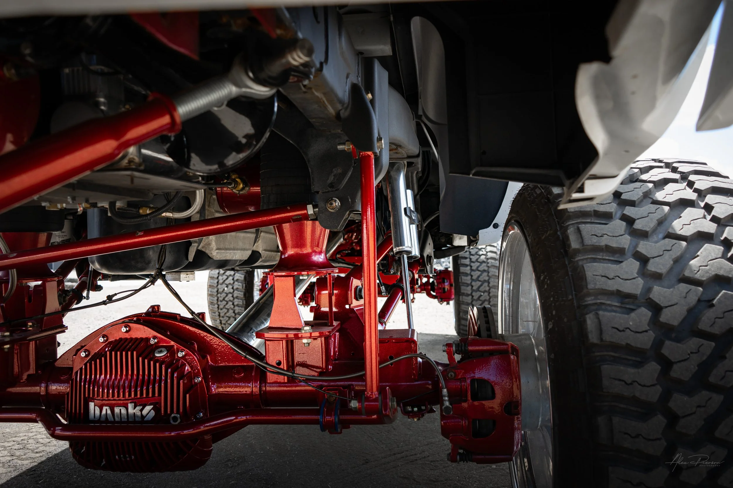 Extensive undercarriage view of a massive Kelderman air suspension lift on a Ram 2500, showcasing red powder-coated links, Fox shocks, and a custom Banks differential cover.
 Lifted truck undercarriage, Kelderman suspension setup, Banks diff cover, R