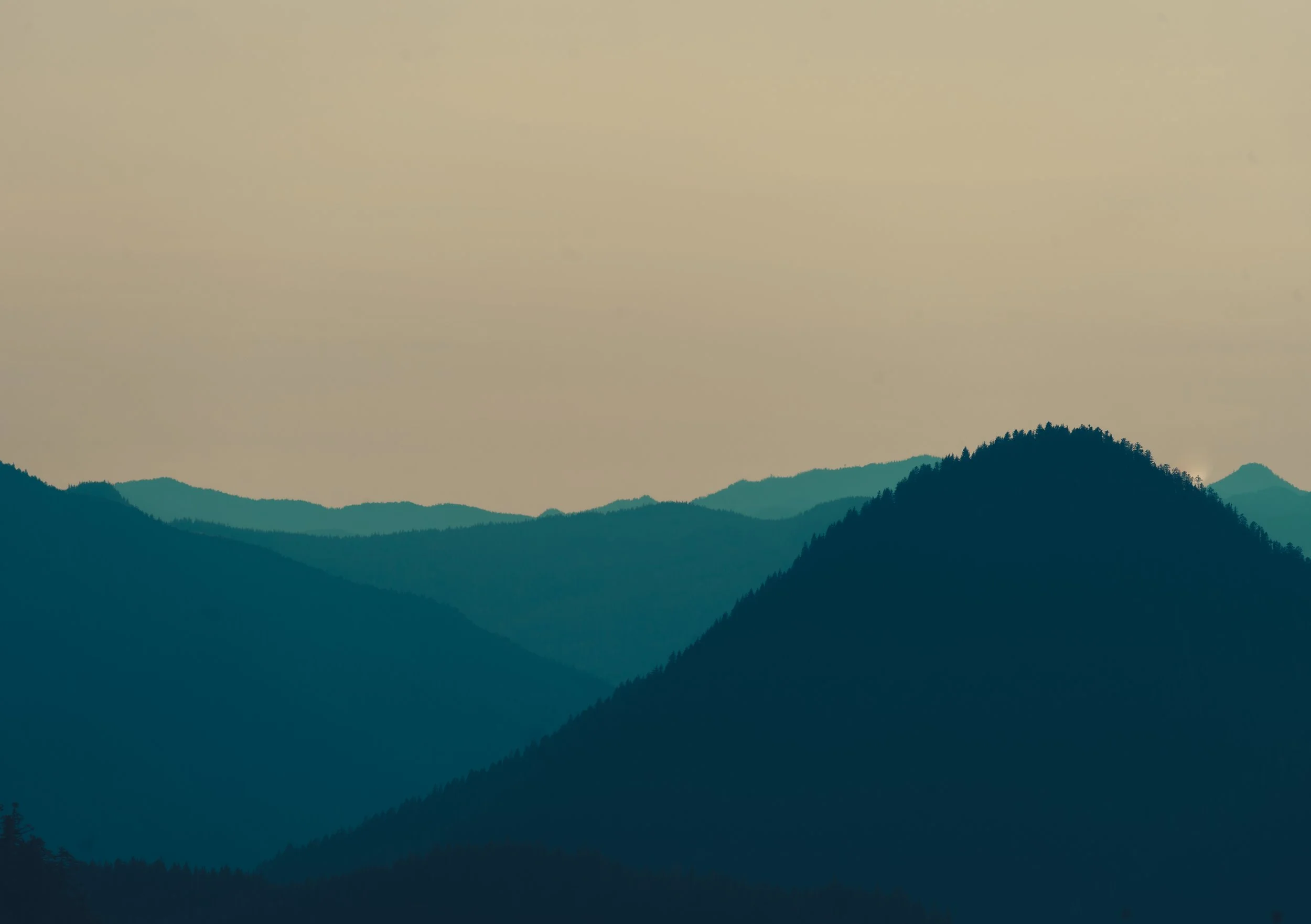 Minimalist landscape photography showing layered blue silhouettes of mountain ridges fading into a hazy, colorful sunset sky in the Pacific Northwest.