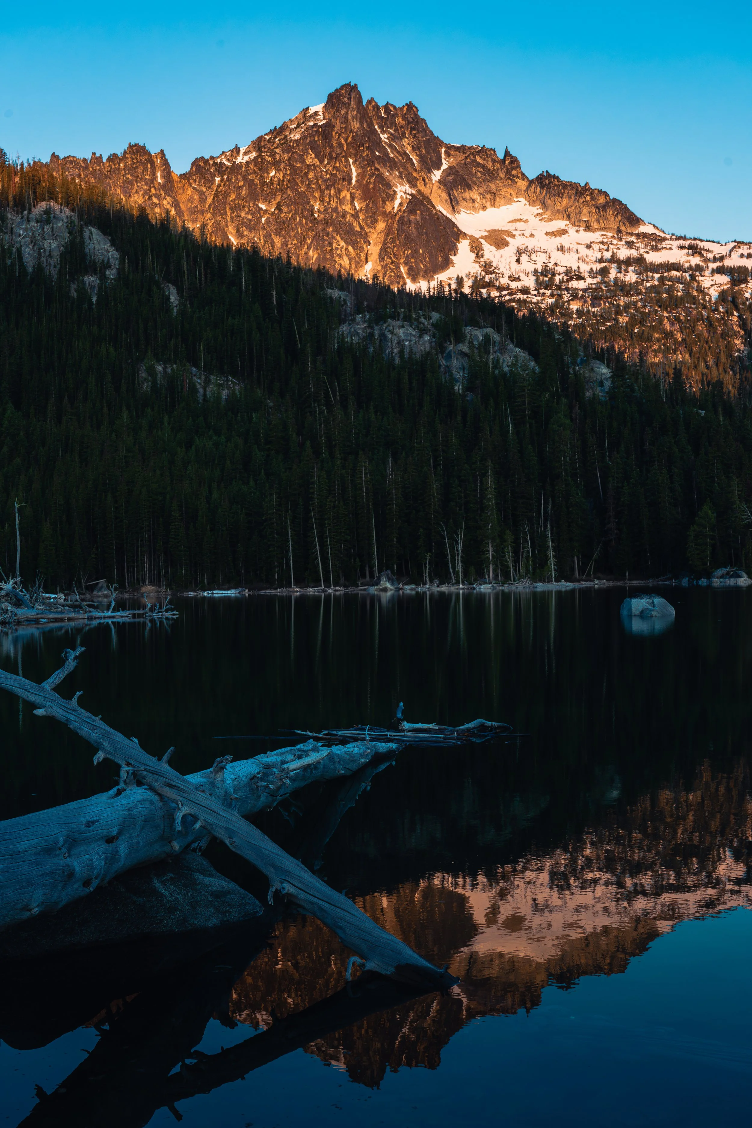 Stunning golden sunrise light hitting a rugged mountain peak and reflecting perfectly in a still, dark alpine lake in the Pacific Northwest – fine art landscape photography.
