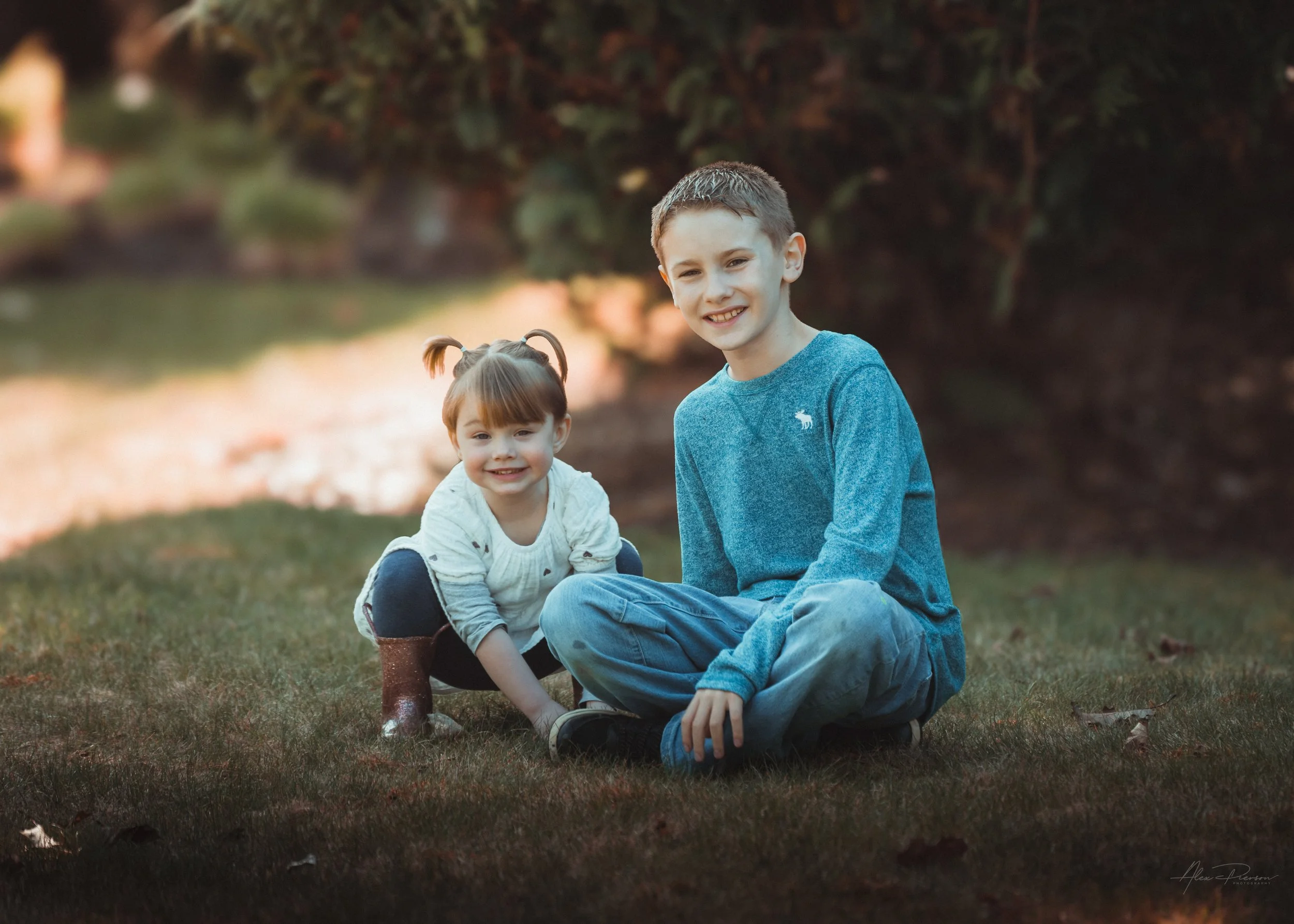 Little girl in a white dress kneeling down in the grass with her cousin, looking directly at the camera during an outdoor portrait shoot in Tumwater, WA– Pacific Northwest lifestyle family and children's photography.