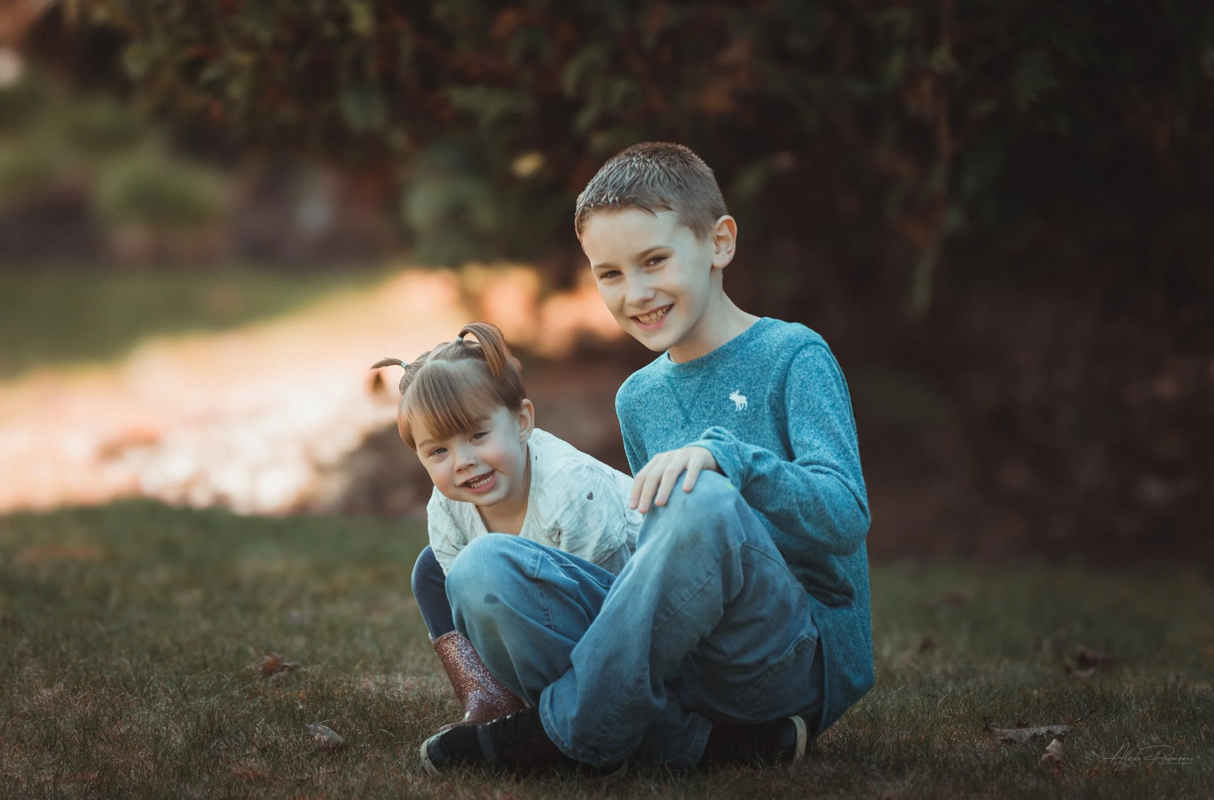 Little girl in a white dress kneeling down in the grass with her cousin, looking directly at the camera during an outdoor portrait shoot in Tumwater, WA– Pacific Northwest lifestyle family and children's photography.