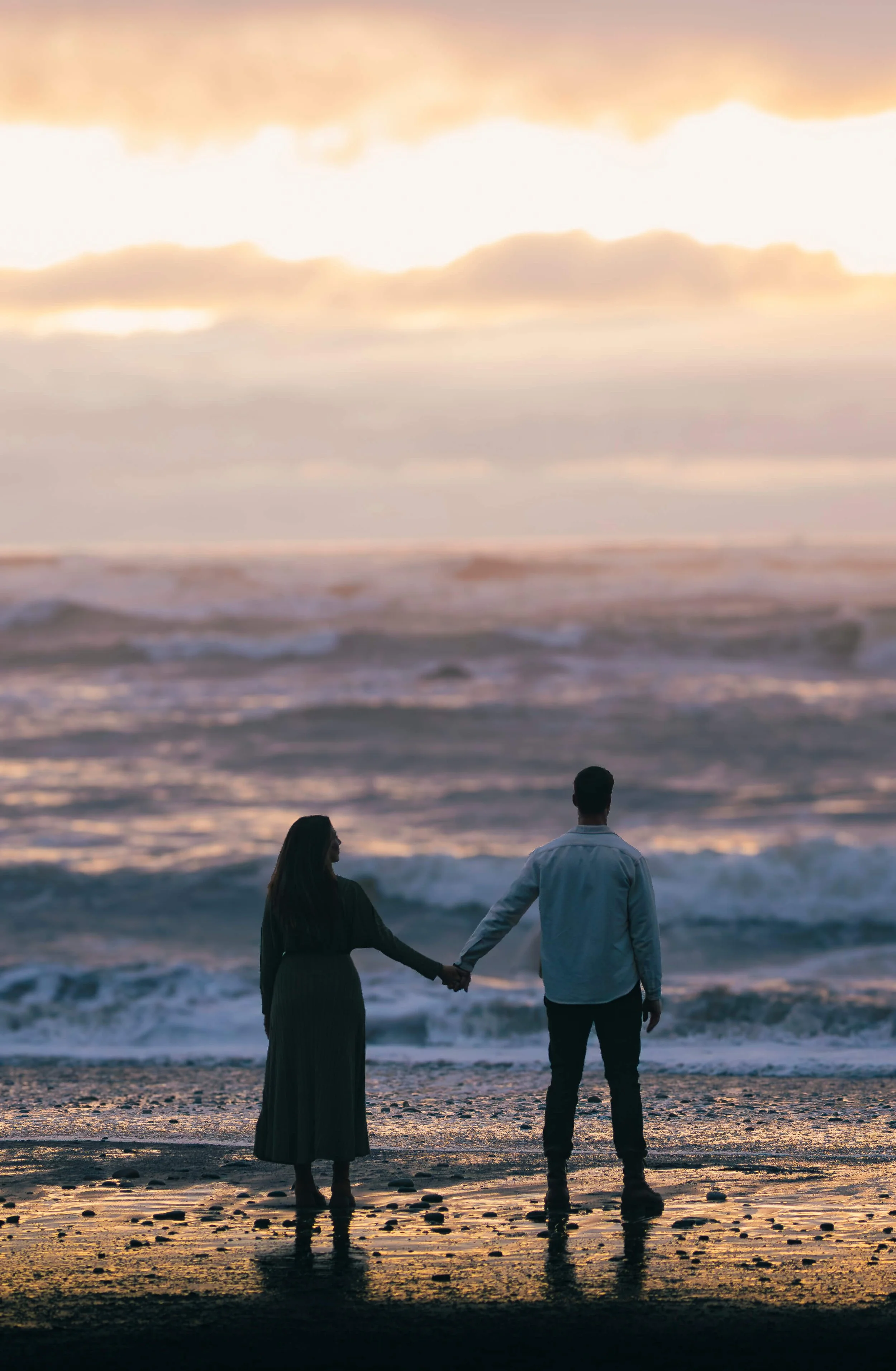 A couple holding hands and looking out over the crashing ocean waves during a romantic sunset at Ruby Beach, WA – Pacific Northwest couples and engagement photography.