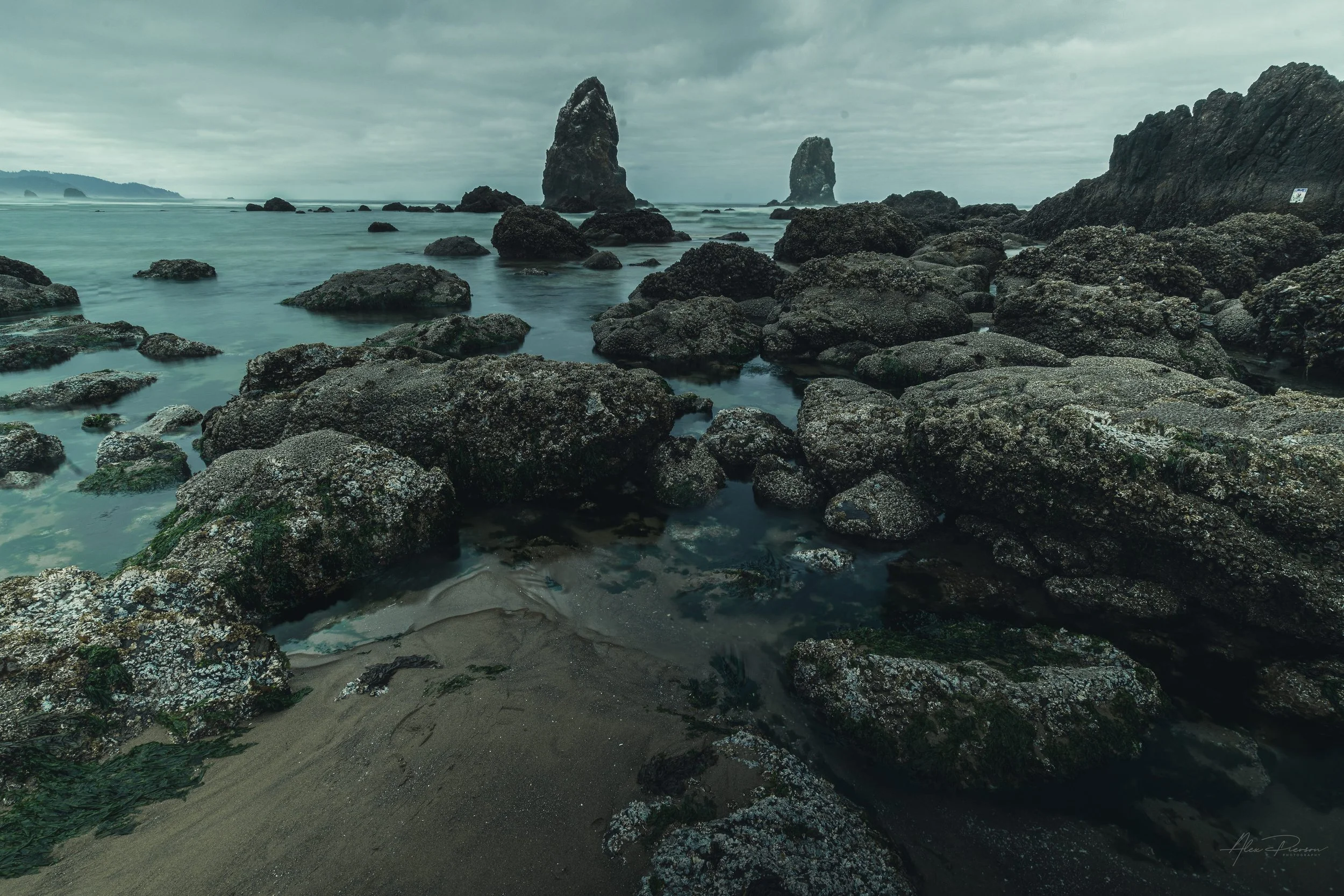 Dark, moody tide pools and rocky shore leading to Haystack Rock under a cloudy sky in Oregon.
The rugged, dark rock formations and calm tide pools of Cannon Beach create a stunning foreground leading the eye out to the iconic Haystack Rock under a mo