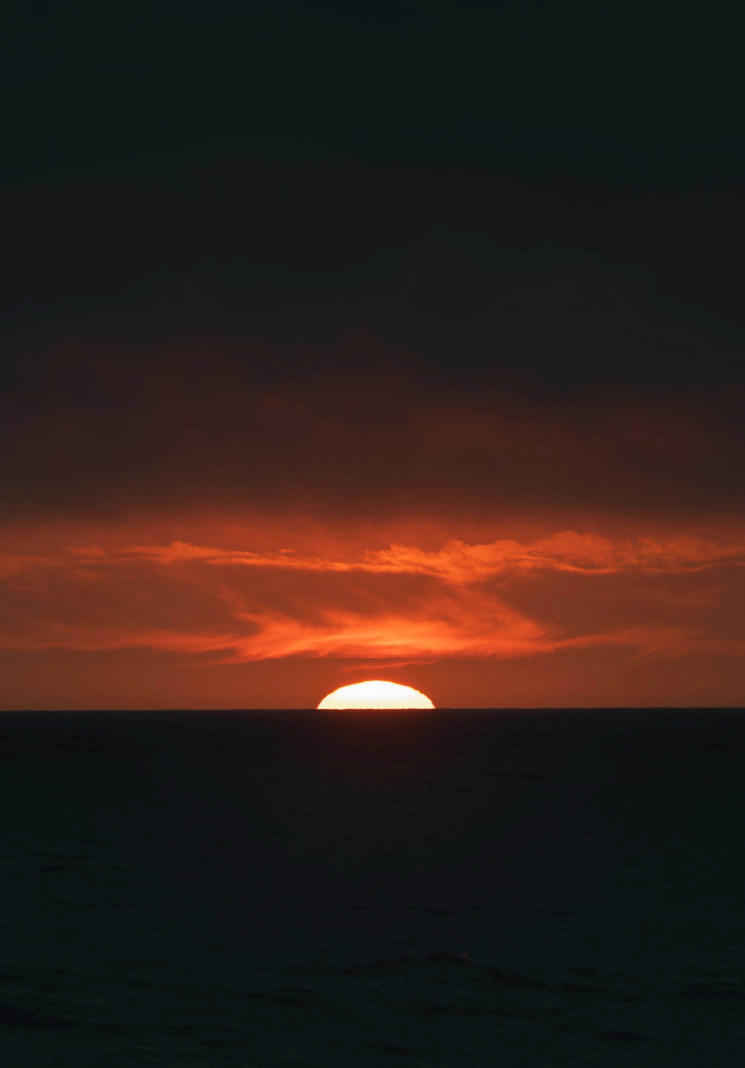 A glowing, fiery red sun half-submerged on the ocean horizon under thick, dark clouds.
The final moments of daylight on the coast. A brilliant, fiery red sun sinks below the ocean horizon beneath a heavy canopy of dark evening clouds.