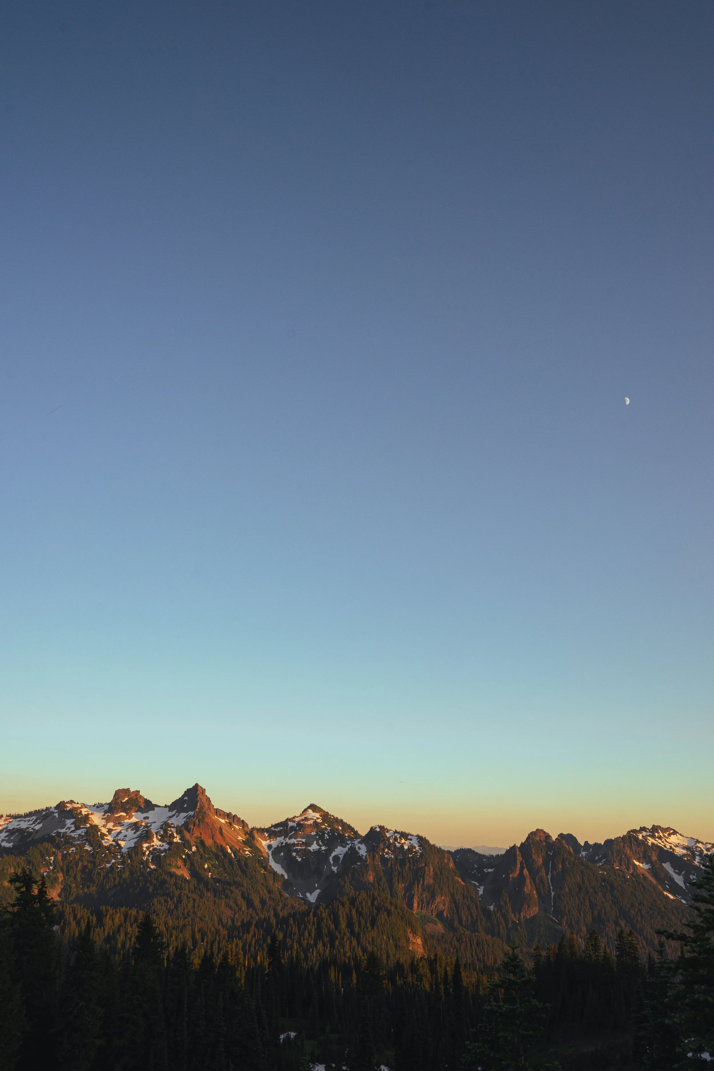 serene twilight view over the jagged peaks of the Tatoosh Range in Mount Rainier National Park, featuring a pale crescent moon in the clear evening sky.