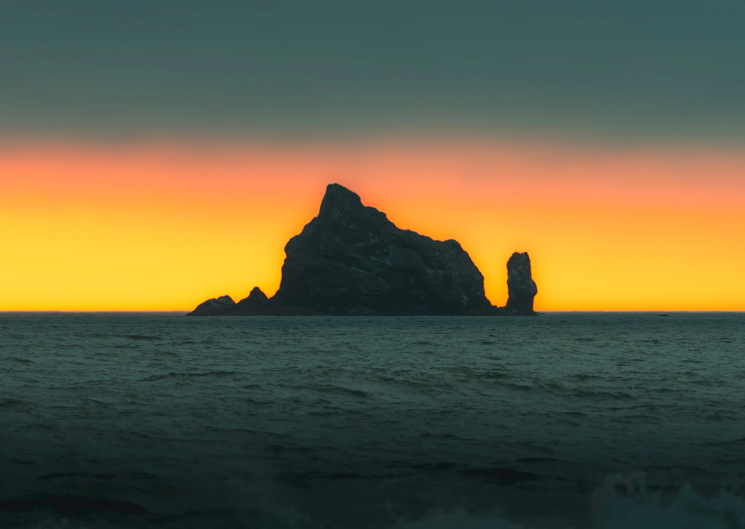 A massive, rocky sea stack silhouetted against a vibrant orange and teal sunset over the ocean in the Pacific Northwest.
The iconic, towering sea stacks of the Pacific Northwest coast stand in stark silhouette against a brilliant, gradient orange and
