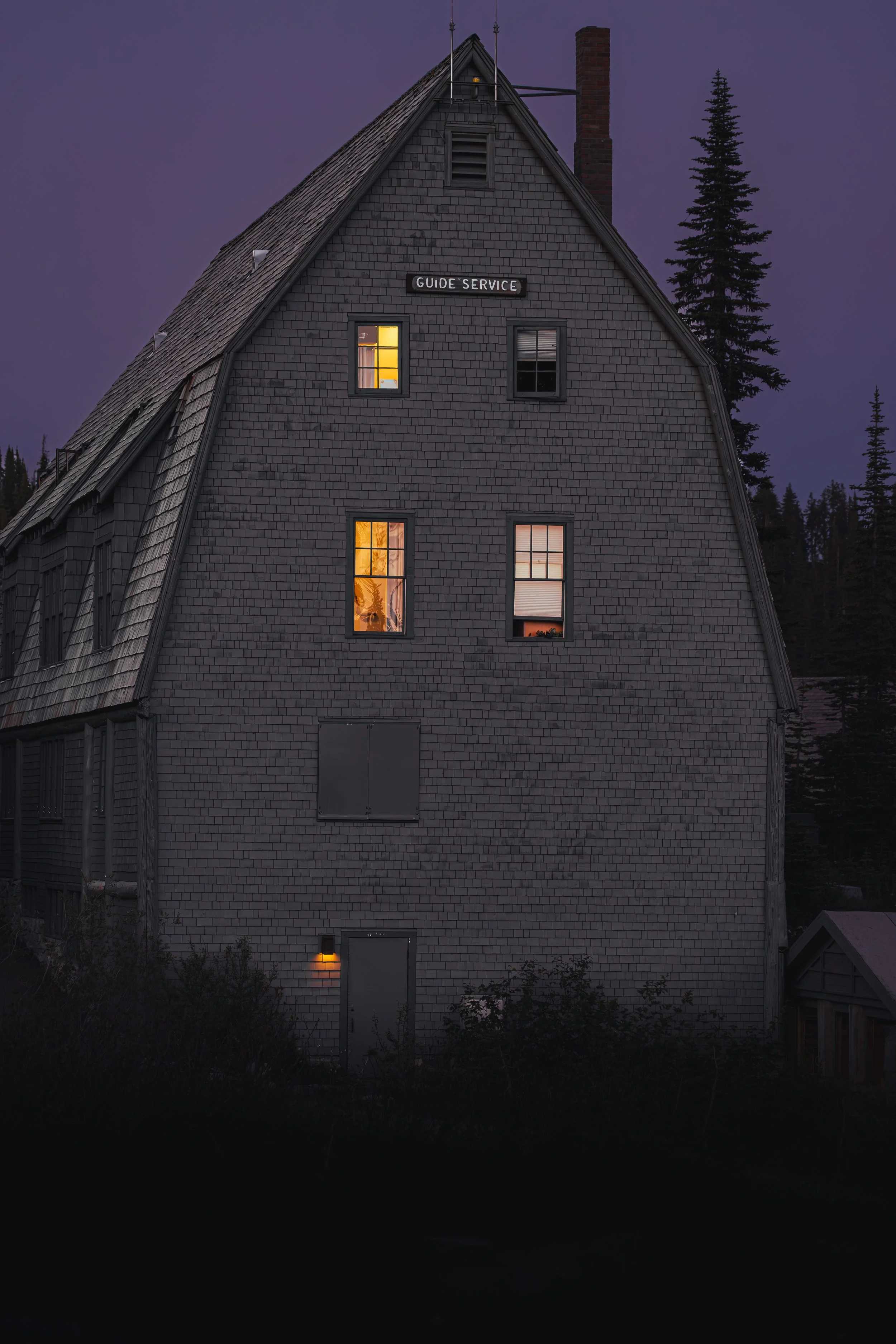Historic shingled Guide Service building with glowing windows under a purple twilight sky at Mt. Rainier.
The historic, shingle-sided Guide Service building at Mount Rainier glows warmly against a striking deep purple twilight sky in the high alpine 