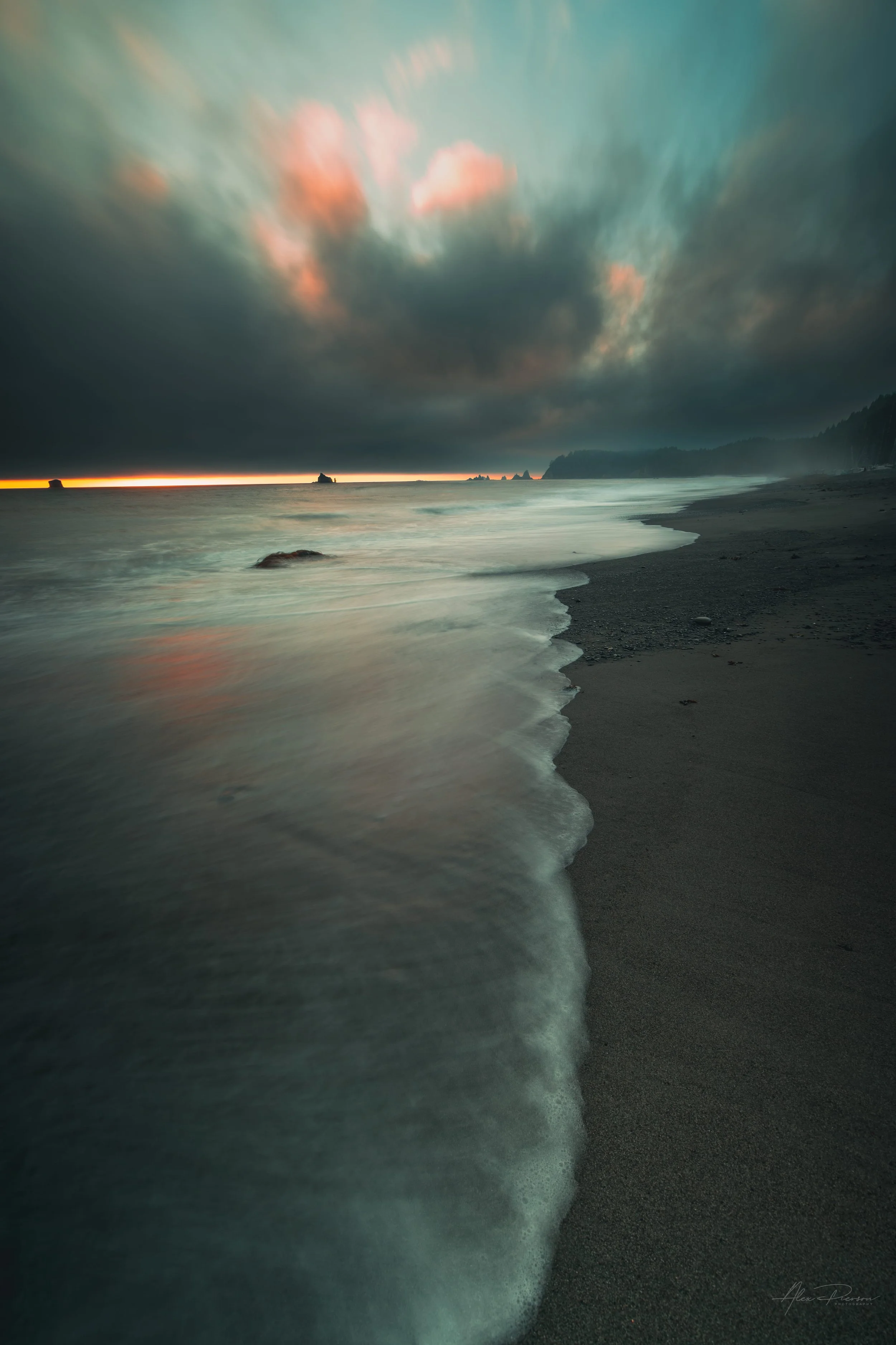 Long exposure photography of waves washing onto a dark sand beach under a moody, cloudy sunset sky with sea stacks in the distance.
A moody, long-exposure capture of the rugged Washington coast. Soft, sweeping waves wash over dark sand beneath a dram