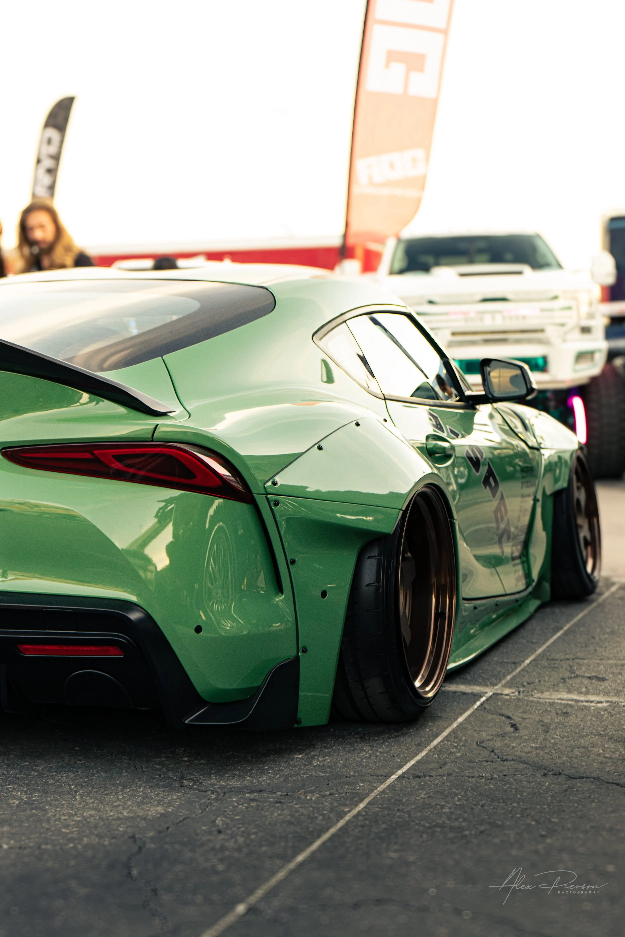 Rear quarter view of a slammed green Toyota Supra showing massive widebody fenders and deep-dish bronze wheels.
 Perfect fitment. A low-angle shot highlighting the massive bolt-on widebody fenders and deep-dish bronze wheels on this custom green MK5 