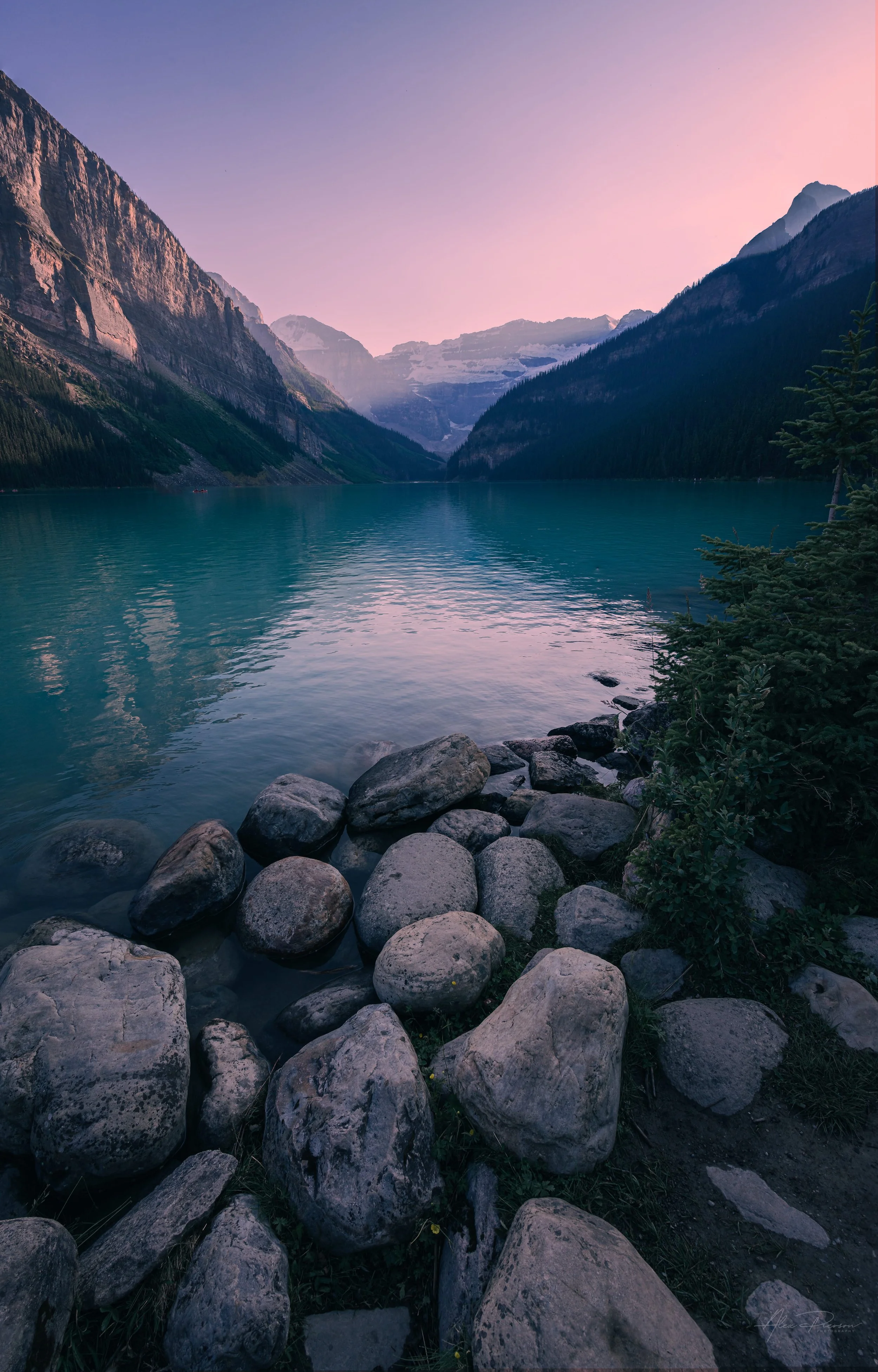Smooth grey rocks line the shore of Lake Louise looking toward a distant glacier under a purple twilight sky.
Smooth, rounded shoreline rocks provide a textured foreground to the calm turquoise waters and distant glaciers of Lake Louise under a stunn