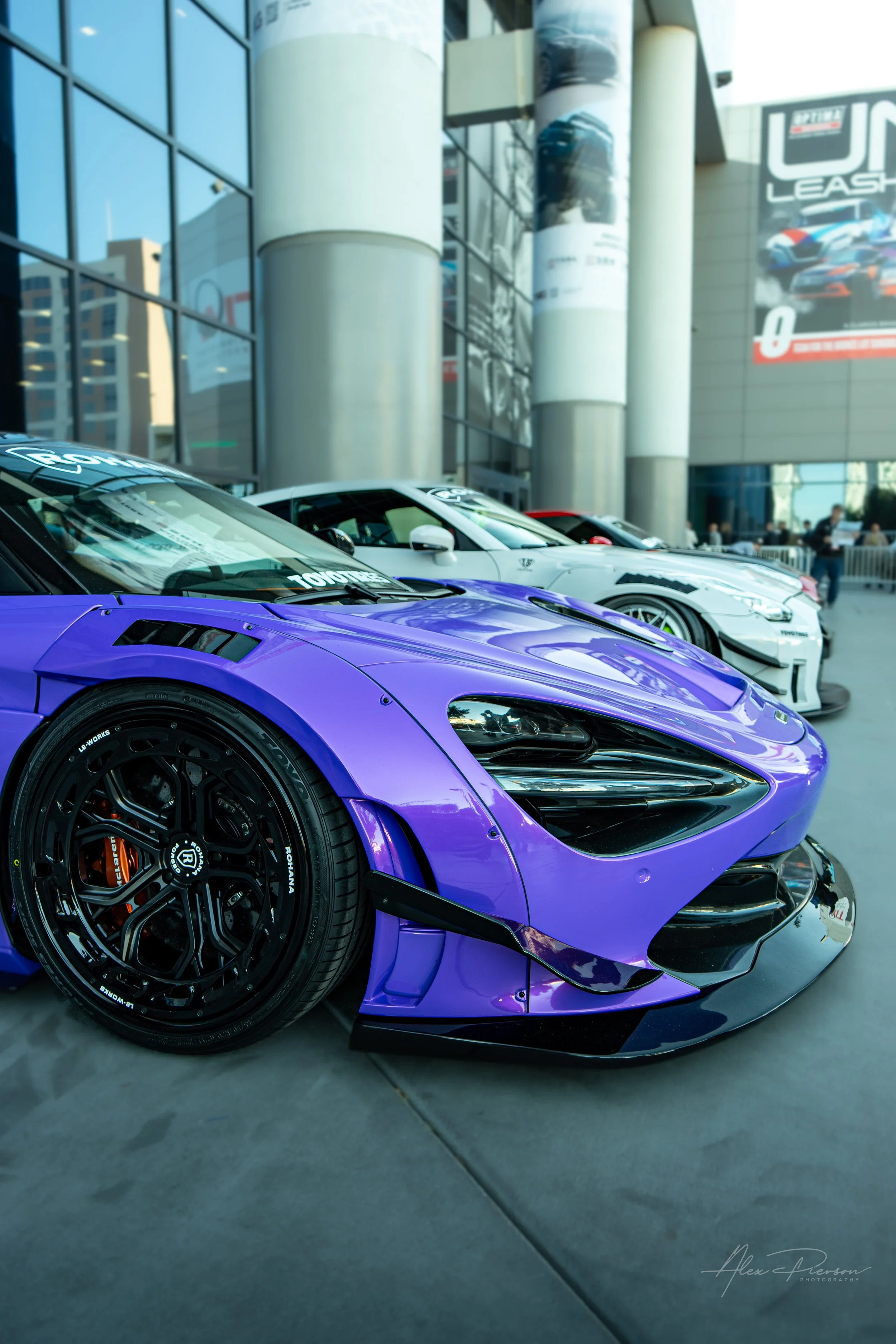 Front angle of a custom purple Liberty Walk widebody McLaren supercar parked at the SEMA Show in Las Vegas.
A striking purple McLaren featuring an aggressive Liberty Walk widebody kit and custom black aftermarket wheels on display outside the SEMA Sh