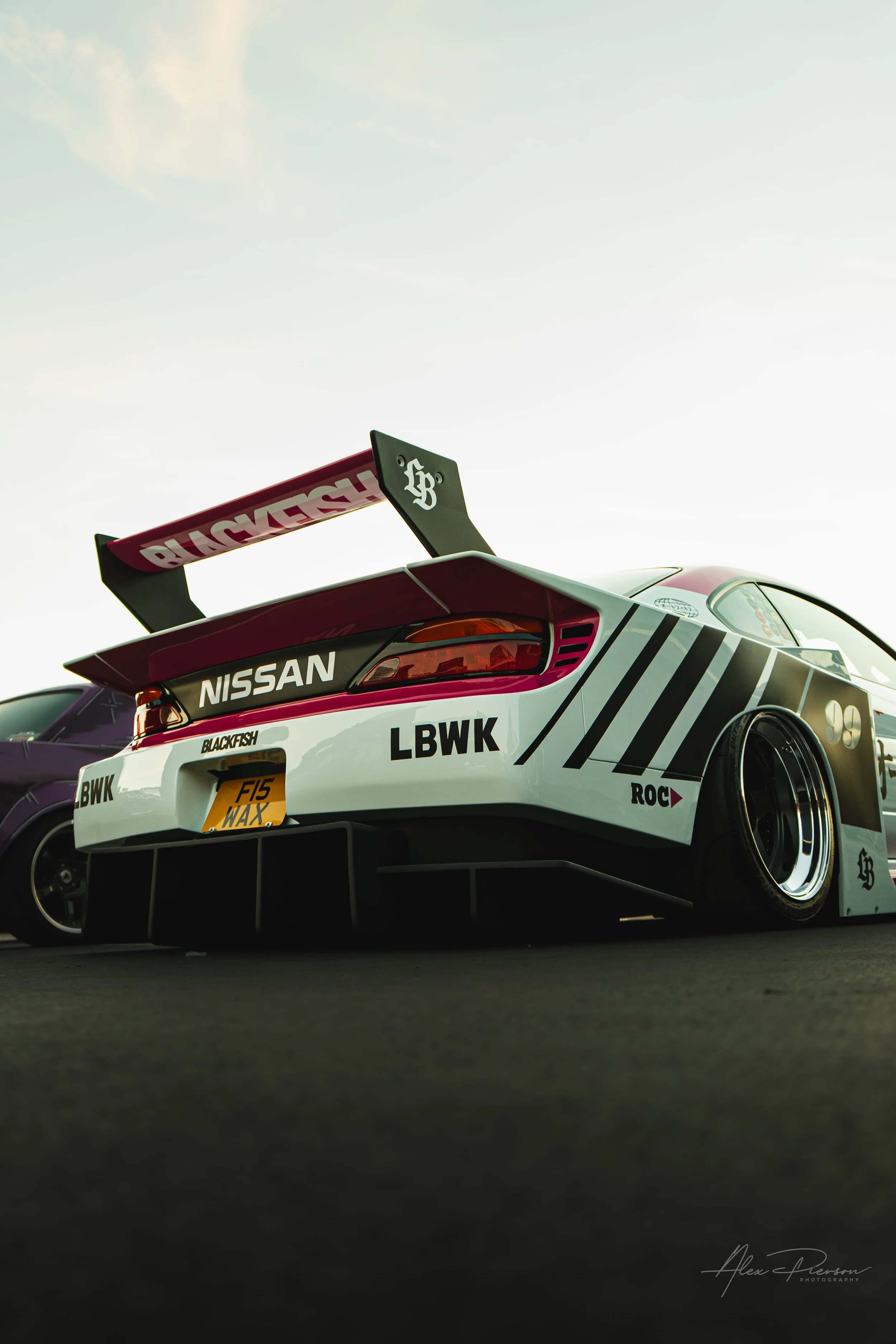 Low angle rear view of a white and pink Liberty Walk Nissan S15 Silvia with a massive racing wing at SEMA.
A ground-level perspective highlighting the aggressive rear diffuser and massive "Blackfish" racing wing on this Liberty Walk Nissan S15 Silvia