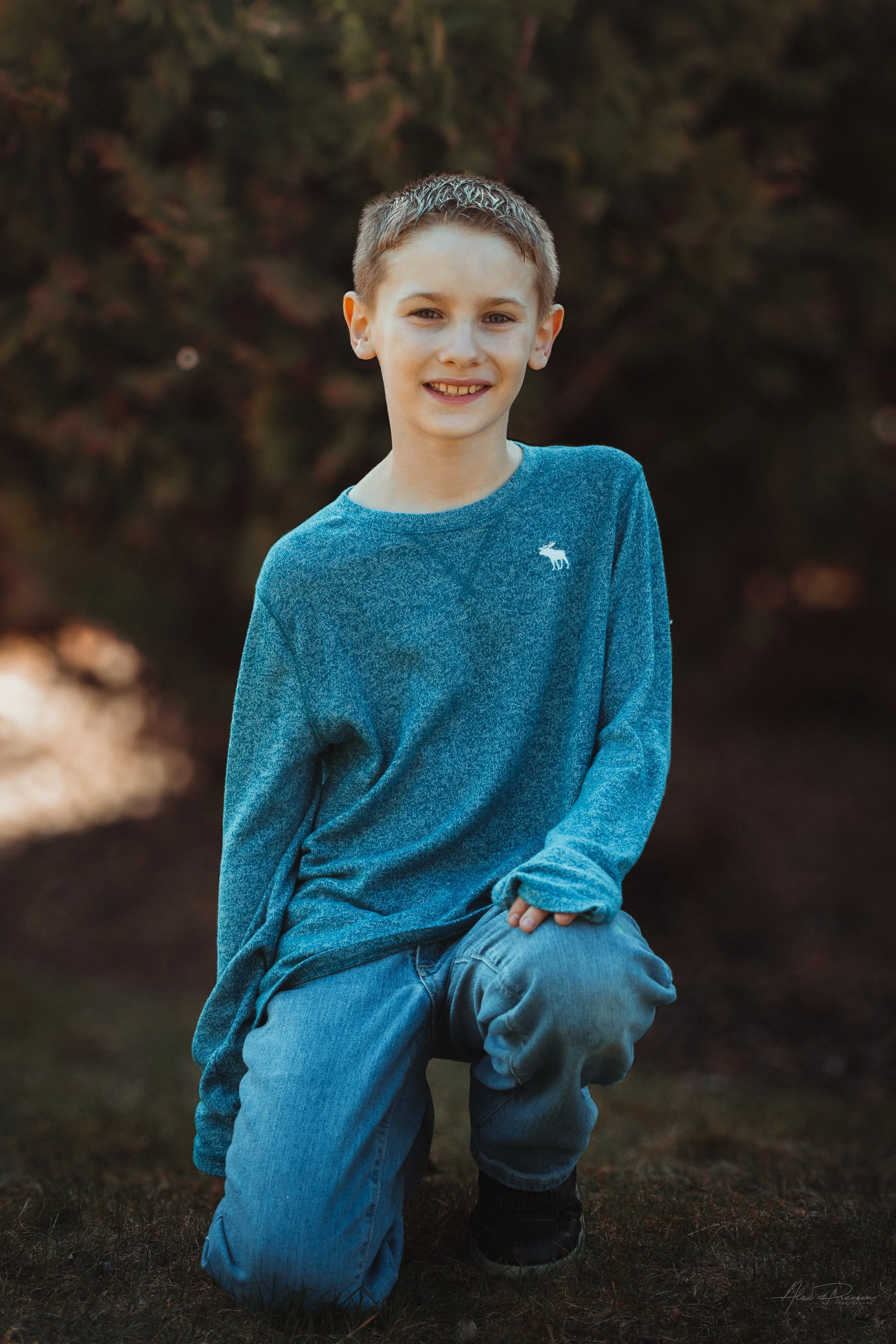 Warm natural light portrait of a young boy smiling while he kneels on one knee while outdoors in Tumwater, WA – Pacific Northwest lifestyle family and children's photography.