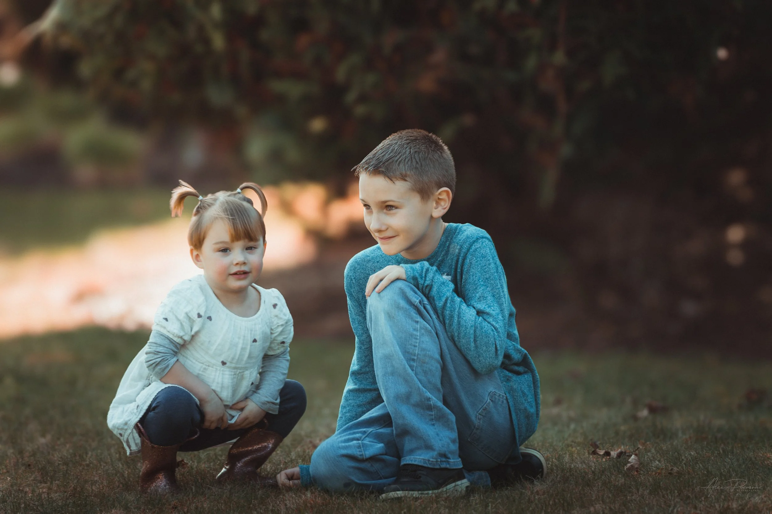 Little girl in a white dress kneeling down in the grass with her cousin, looking directly at the camera during an outdoor portrait shoot in Tumwater, WA– Pacific Northwest lifestyle family and children's photography.