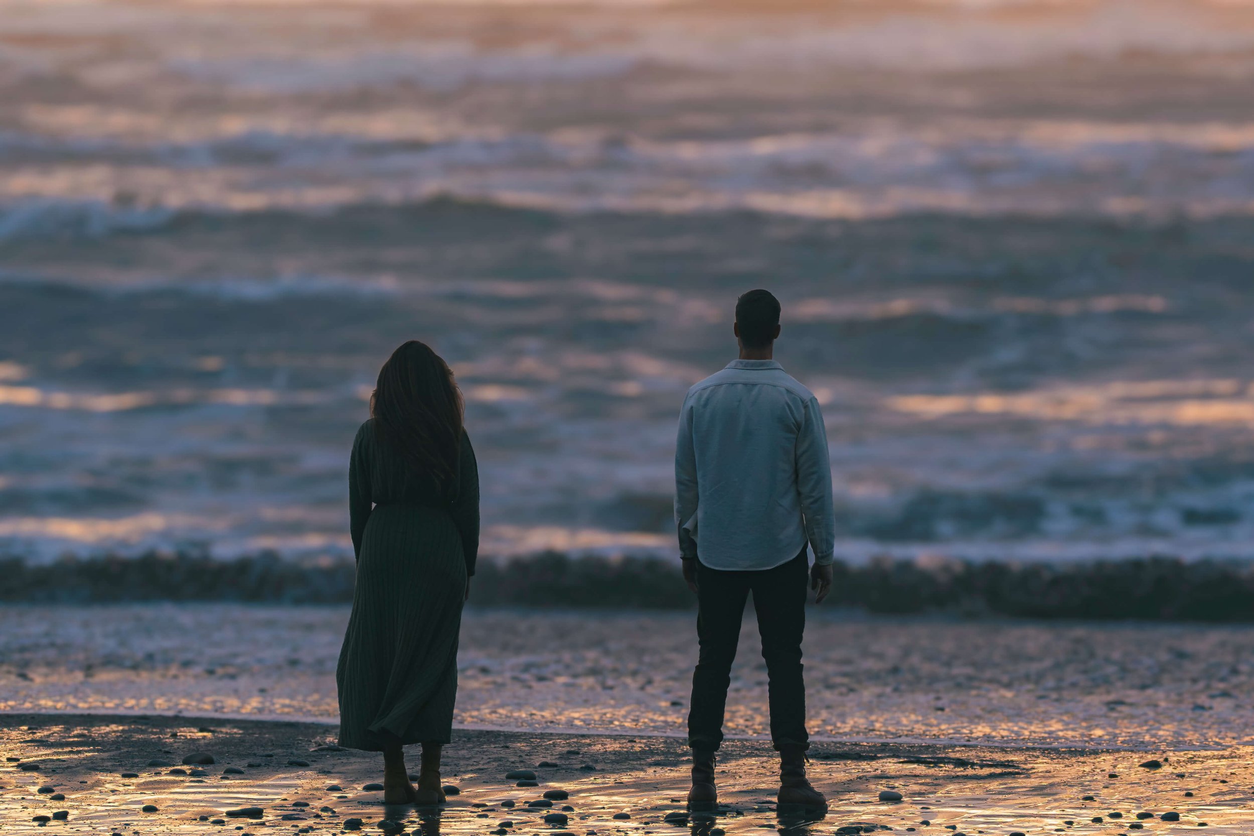 A man and woman standing on the beach watching the moody ocean waves roll in during twilight at Ruby Beach, Washington – Pacific Northwest coastal portrait photography.