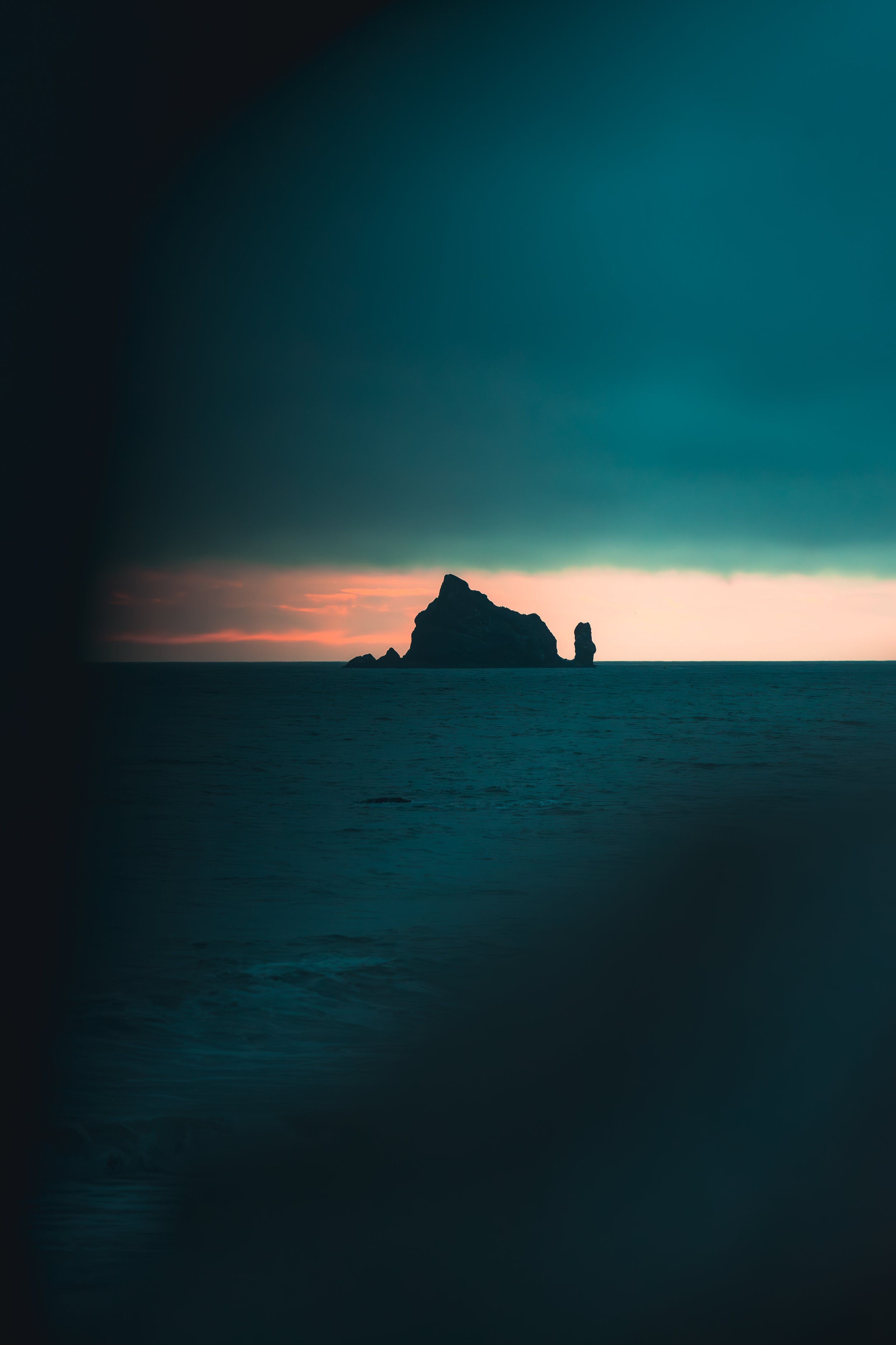 A distant sea stack silhouetted against a narrow band of sunset light, surrounded by deep, moody teal storm clouds and dark ocean.
Dark, moody, and atmospheric. A solitary sea stack breaks the horizon as a sliver of warm sunset light pierces through 