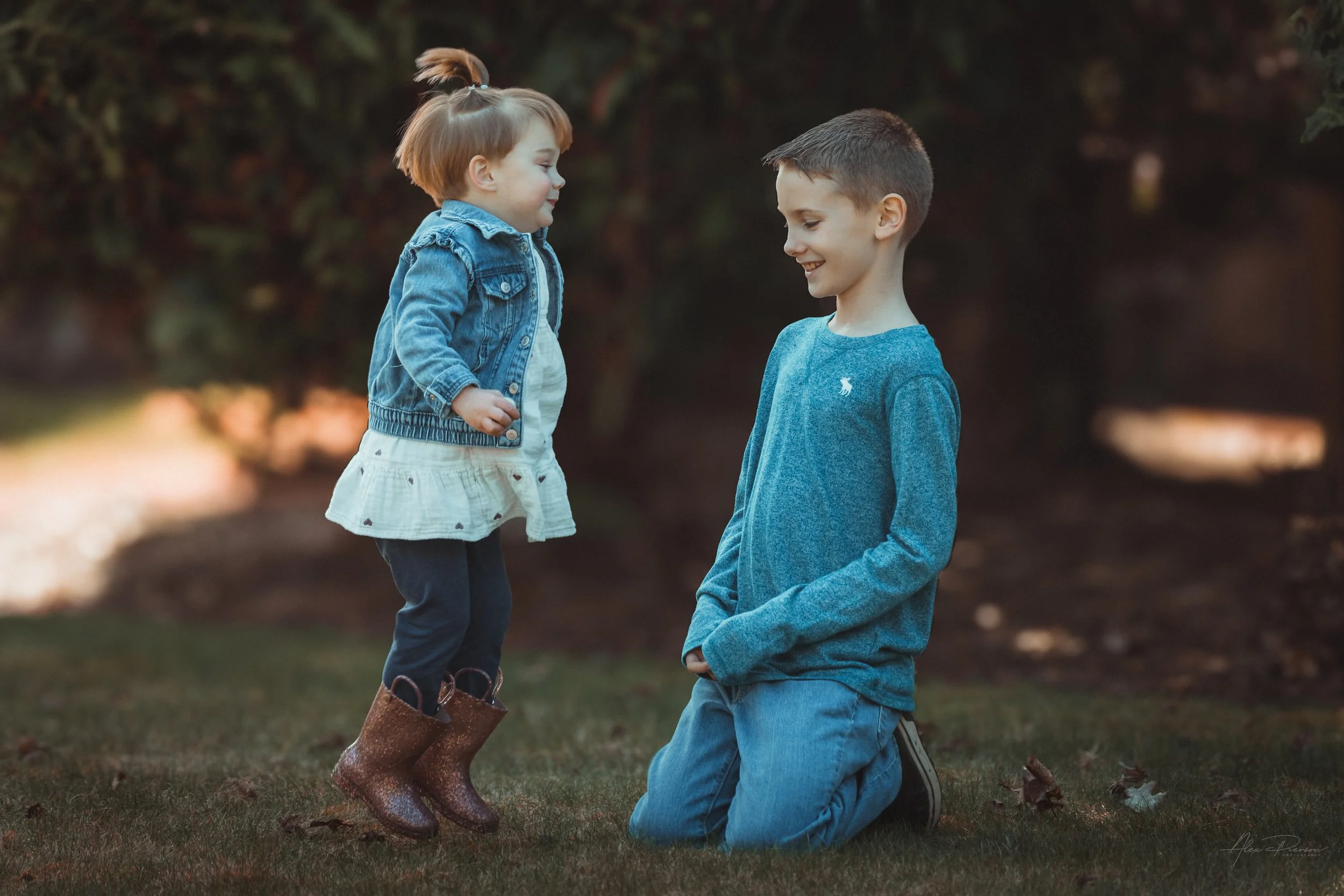 little girl wearing a jean jacket, white dress, and small brown boots jumping off the ground while staring at her cousin during a family photoshoot  in Tumwater, WA – Pacific Northwest lifestyle family and children's photography.