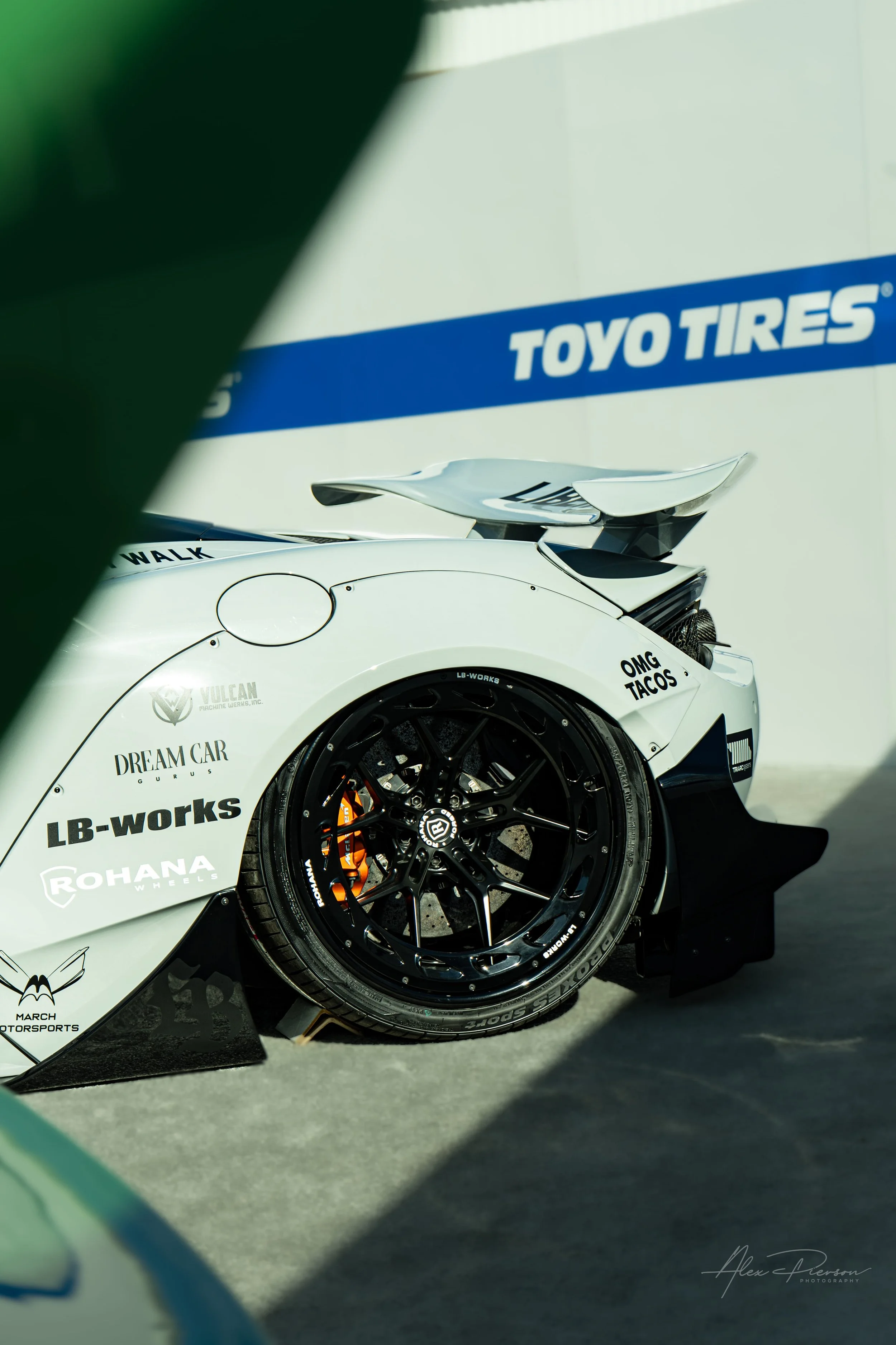 McLaren 720S showing a massive black wheel and aggressive widebody kit.
Supercar stance in the Toyo Tires Treadpass. Framed perfectly by the foreground, this white Liberty Walk McLaren 720S shows off its aggressive rear overfenders, active aero wing,