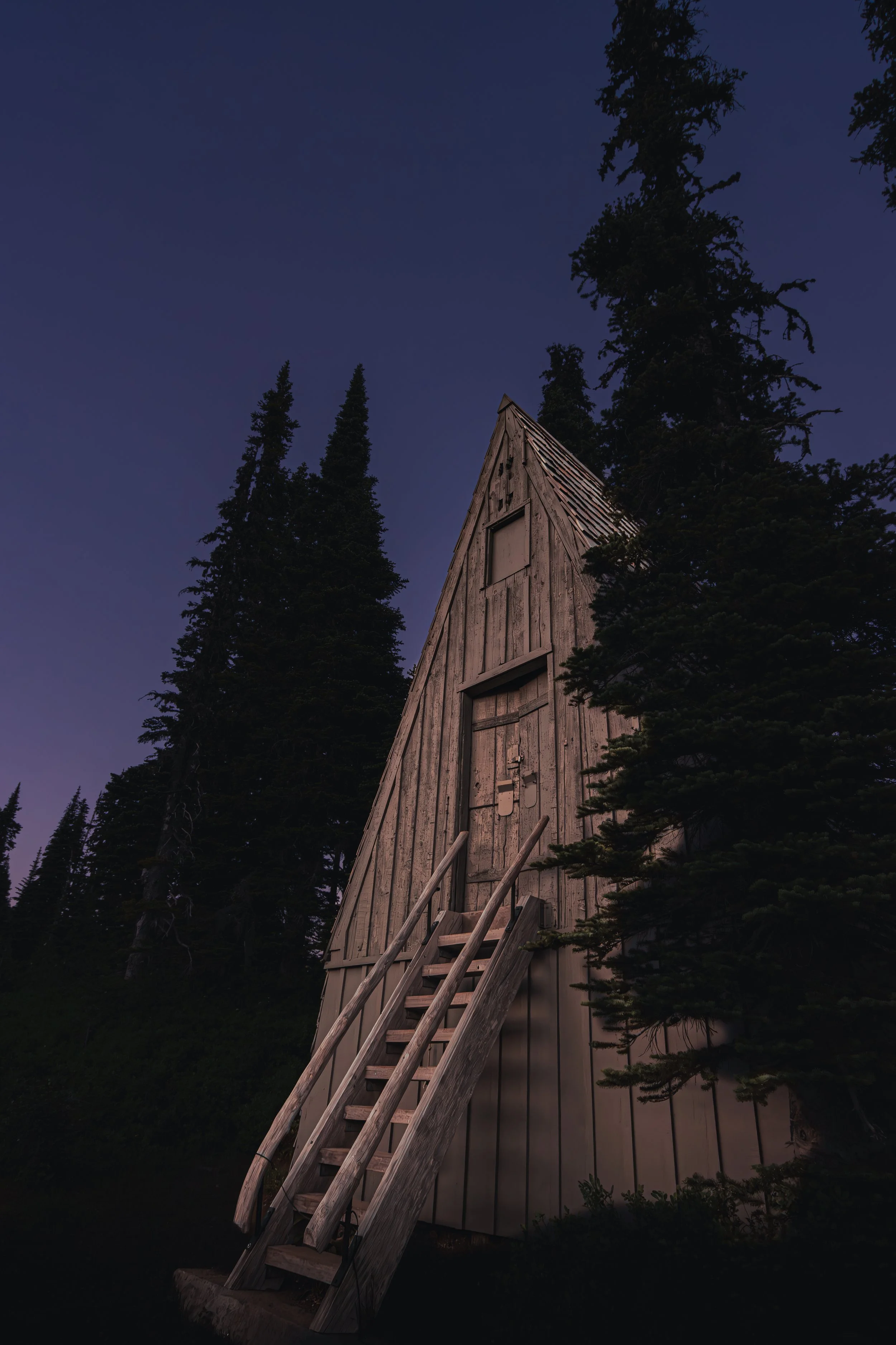 A rustic wooden A-frame cabin tucked in a dark pine forest at twilight in Mt. Rainier National Park.
A classic, weathered A-frame mountain shelter sits quietly among the towering evergreens as night falls over the Mount Rainier high country.