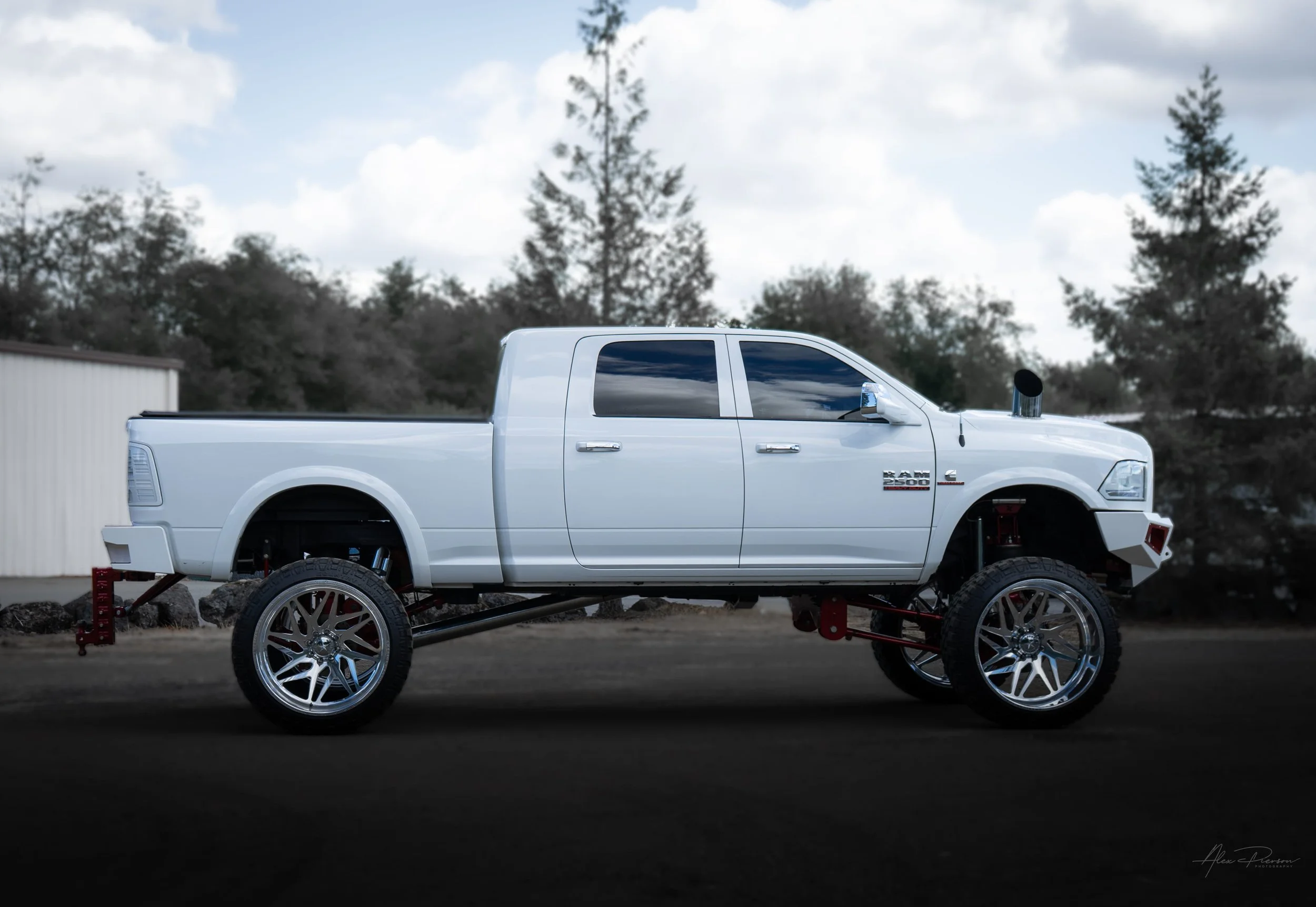 Full side profile of a custom lifted white Dodge Ram 2500 featuring a red Kelderman air suspension lift, custom hood stack exhaust, and polished forged wheels.
Lifted Ram 2500 side profile, Kelderman lift kit, Cummins hood stack, white Ram truck.
whi