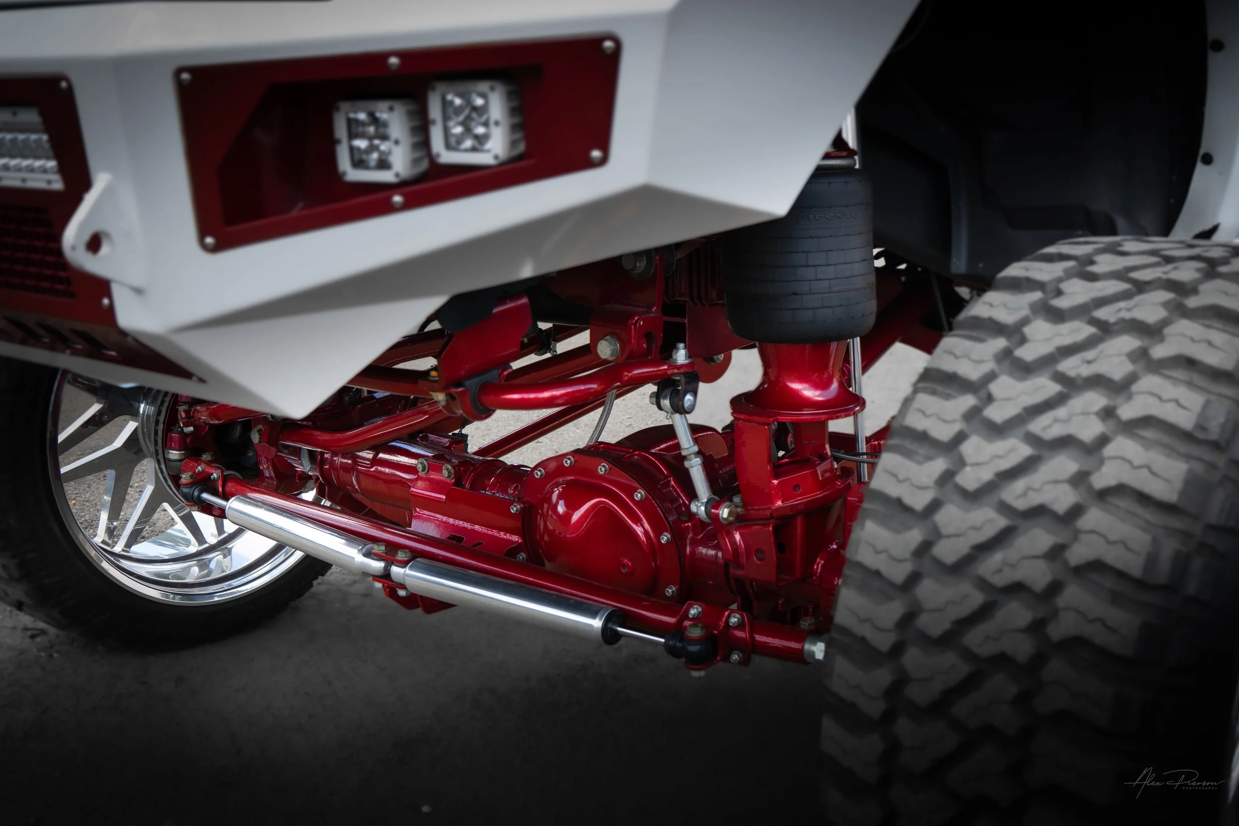 Under-carriage view of a custom Ram truck highlighting a candy red powder-coated front differential, Kelderman air suspension steering components, and a white Bodyguard bumper.
 Red front axle, Kelderman front suspension, Bodyguard front bumper, Ram 