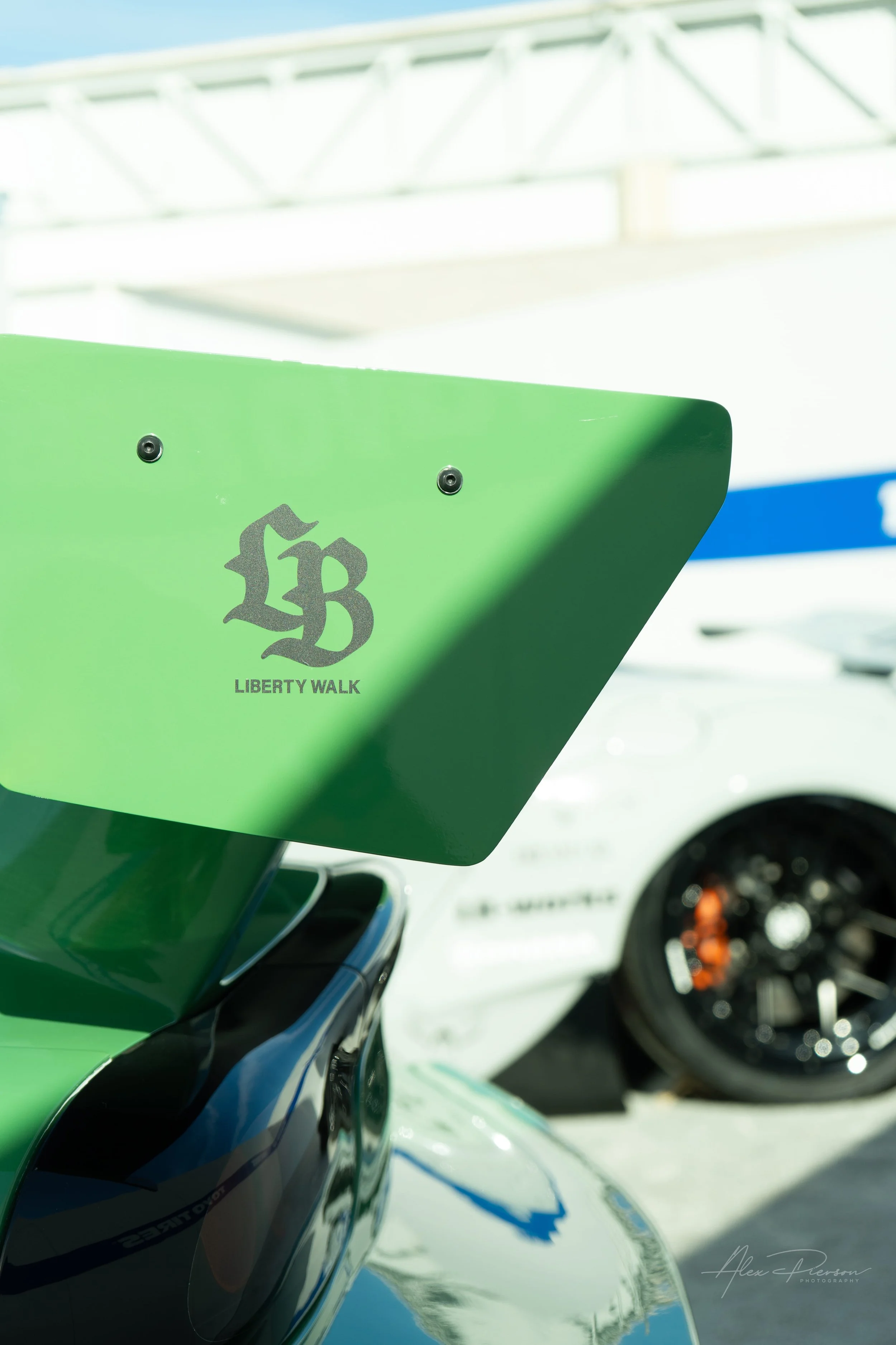 Close up of a green Liberty Walk rear wing endplate with a white widebody McLaren blurred in the background.
Signature aerodynamics. A detailed shot of the classic LB-Works logo on a custom green Mazda rear wing, with a slammed white McLaren lurking 