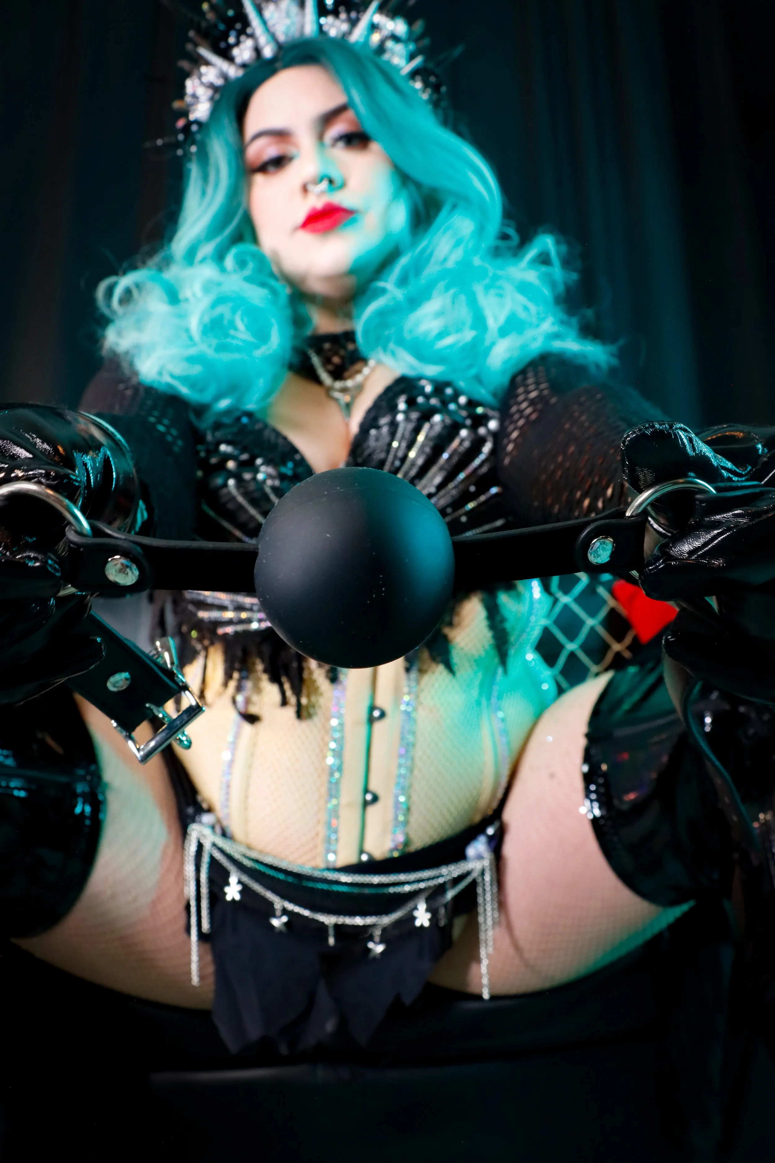 MerDomina stares down at the viewer from a black sofa in shiny black thigh-high heel boots, turquoise hair, and a sparkly black mermaid burlesque-inspired costume, holding a ball gag up to the viewer's mouth