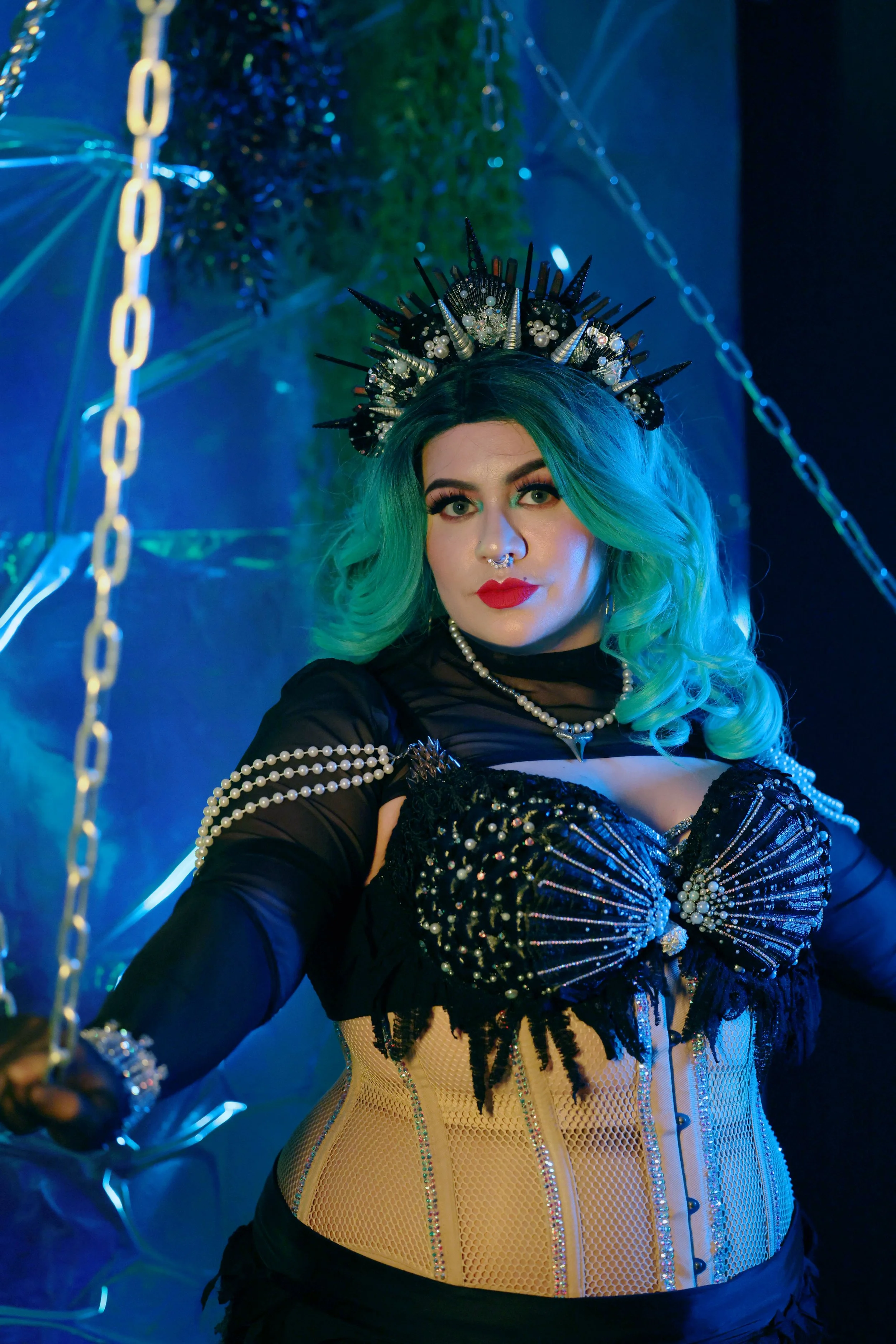 MerDomina holds chains in shiny black thigh-high heel boots, turquoise hair, and a sparkly black mermaid burlesque-inspired costume