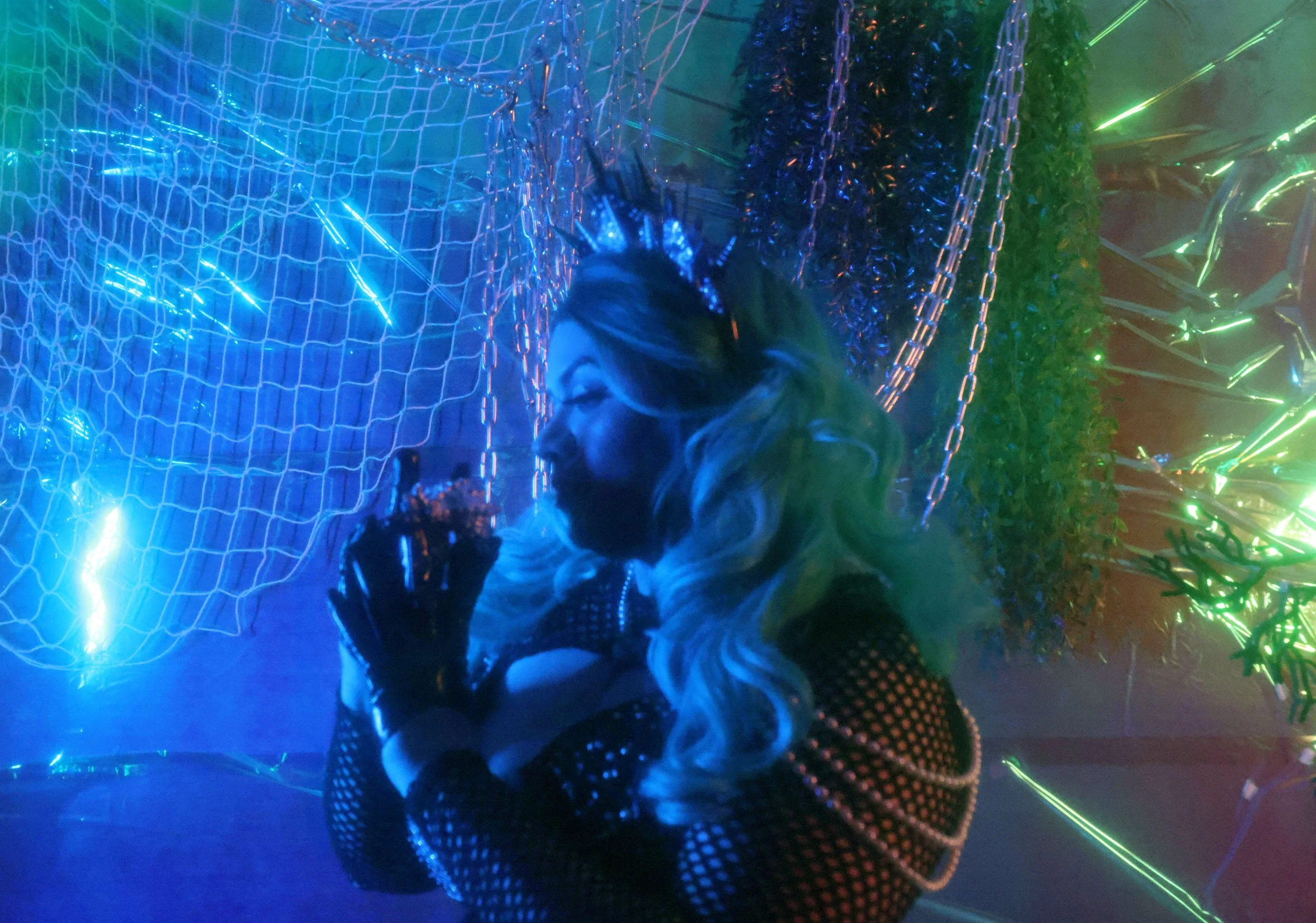 MerDomina looks wistfully at a shell from profile view in multicolored lighting, in turquoise hair and a sparkly black mermaid burlesque-inspired costume
