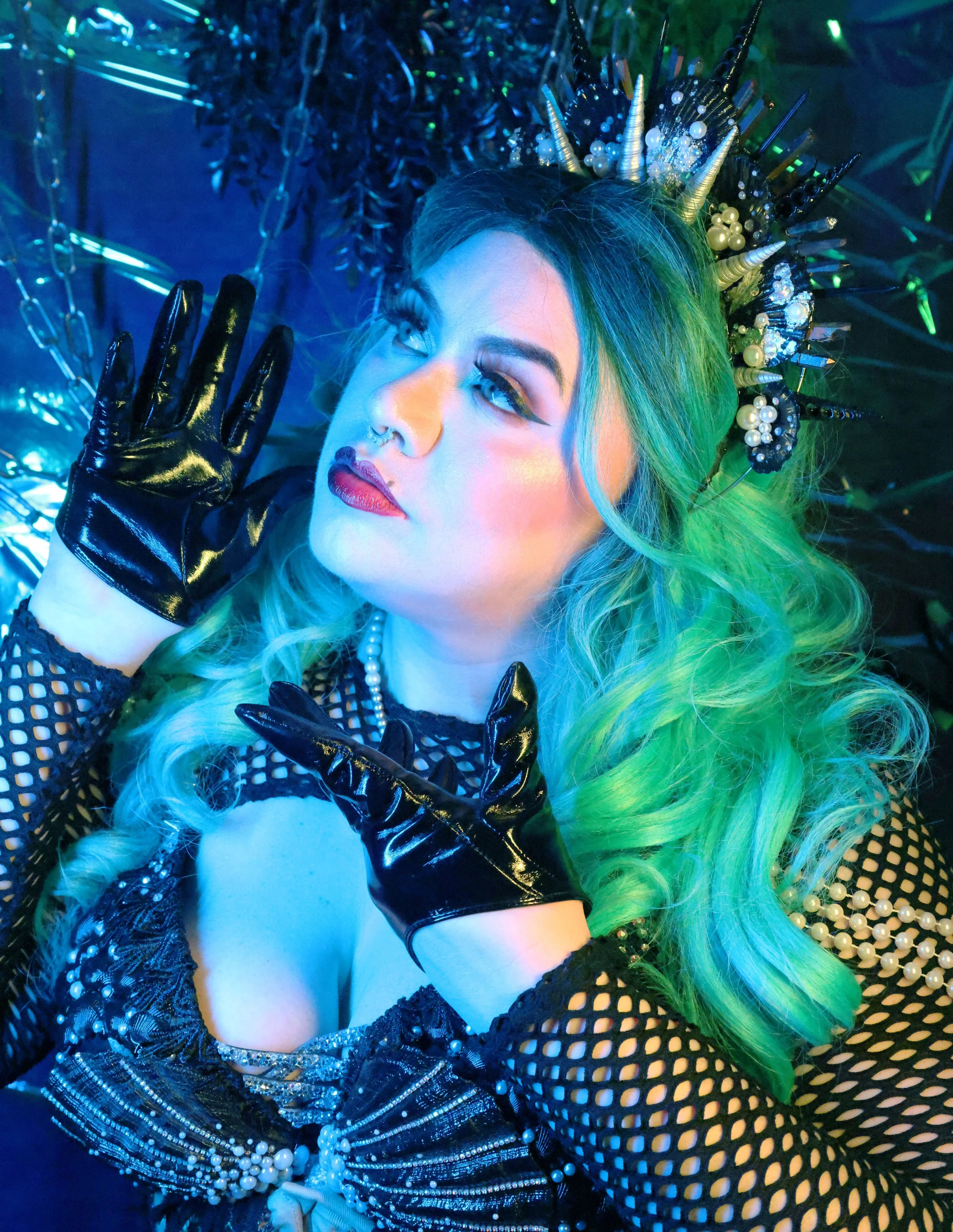 MerDomina playfully frames Her face with PVC-gloved hands in turquoise hair, red lips, and a handmade shell crown