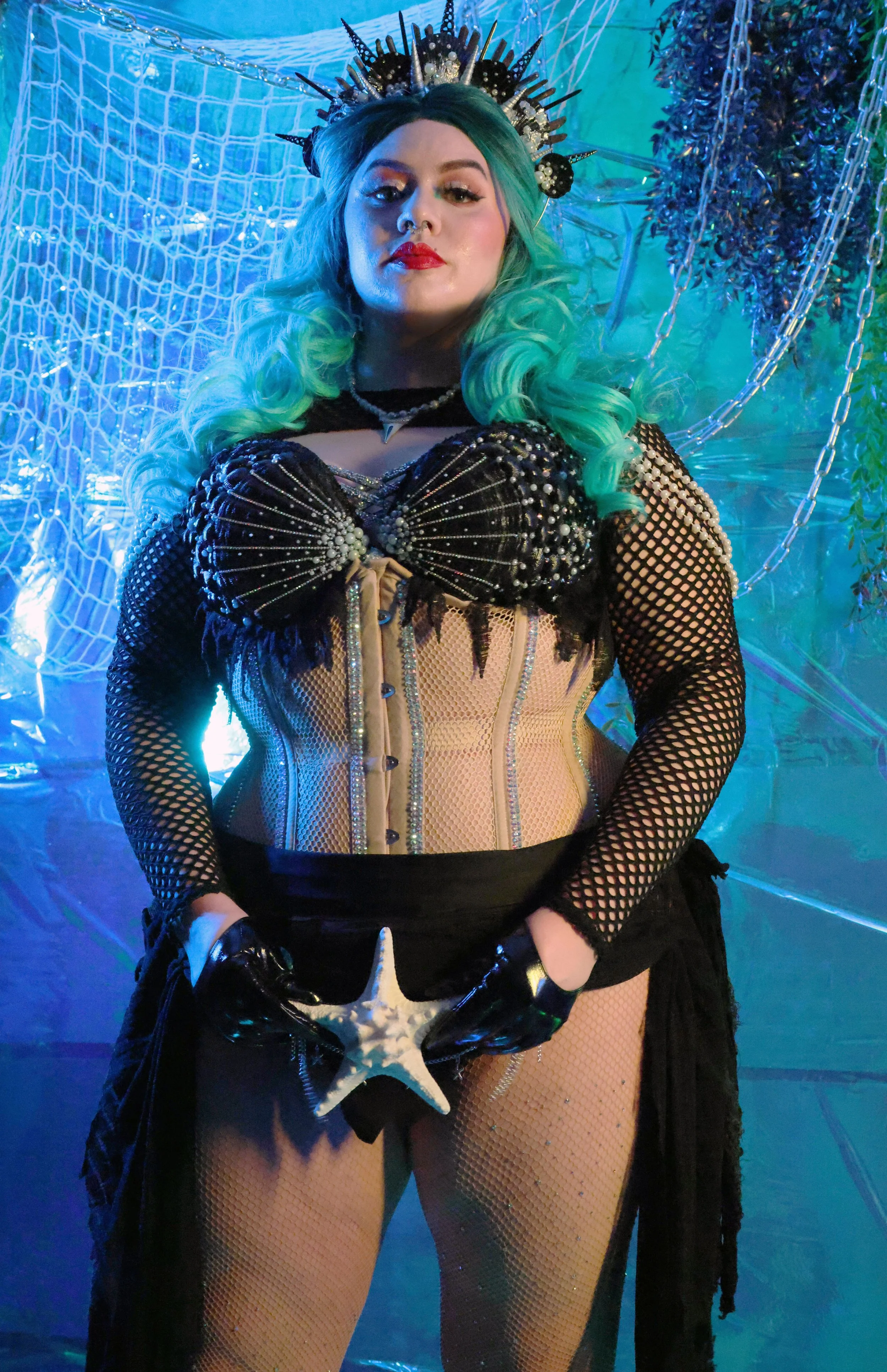 MerDomina poses in turquoise hair and a sparkly black mermaid burlesque-inspired costume with a white starfish in front of the treasure between Her legs