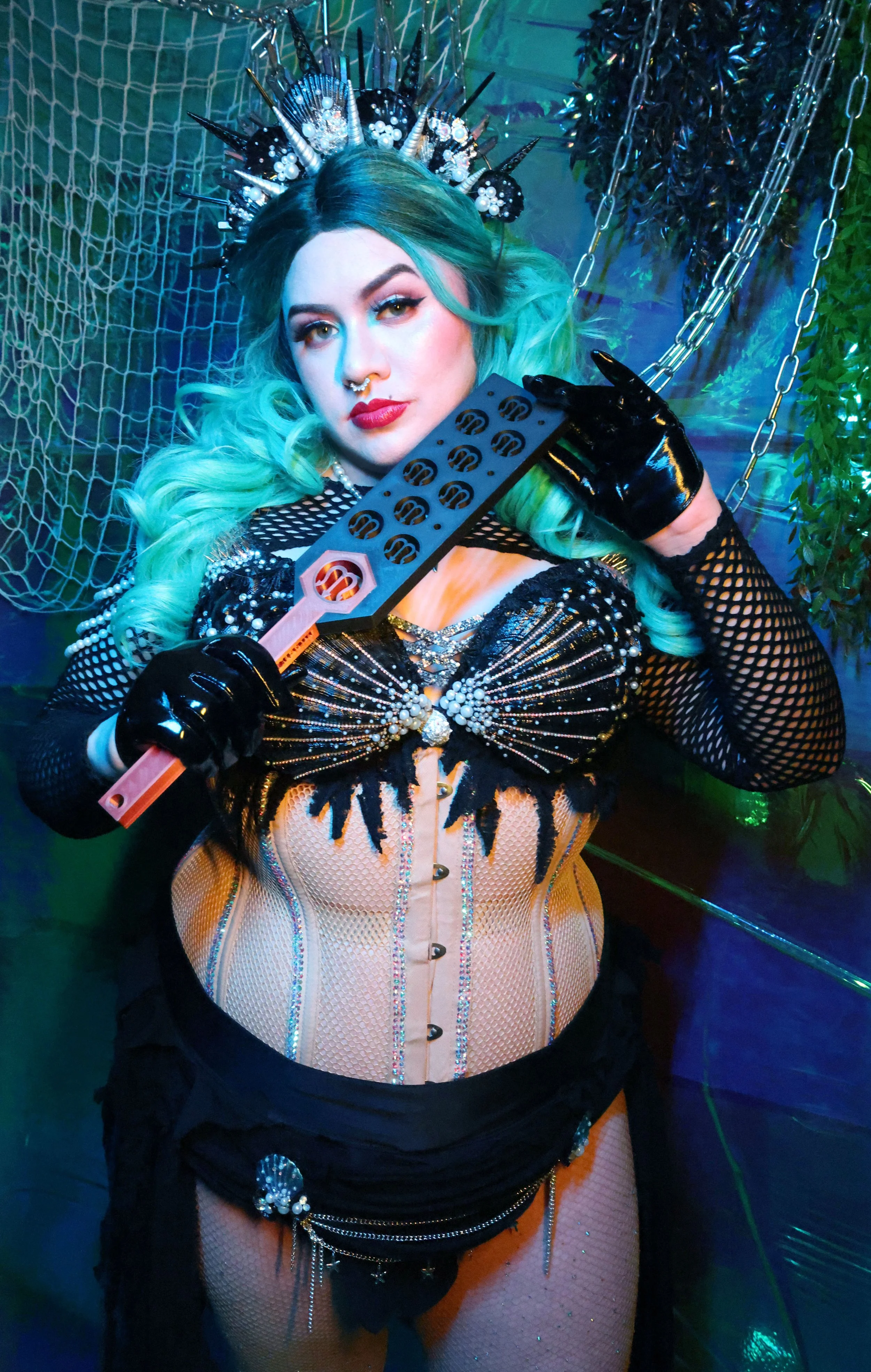 MerDomina holds a black & copper custom logo paddle in turquoise hair and a sparkly black mermaid burleque-inspired costume