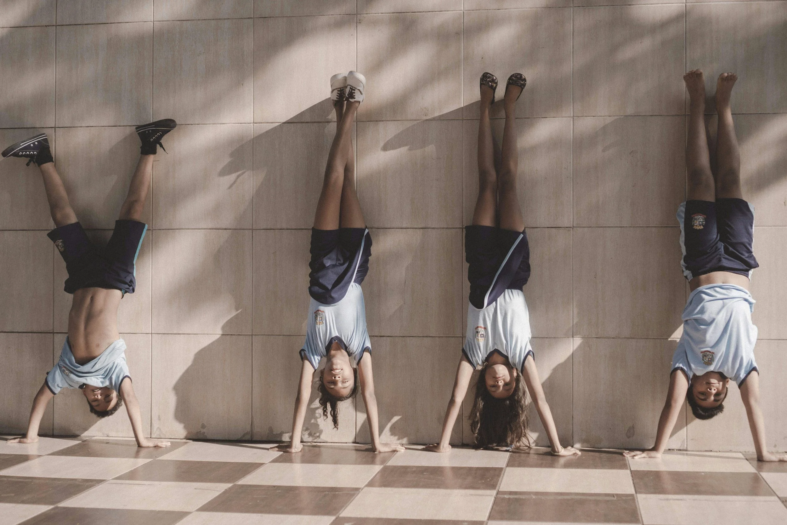 Children in sports uniforms doing handstands against a tiled wall.