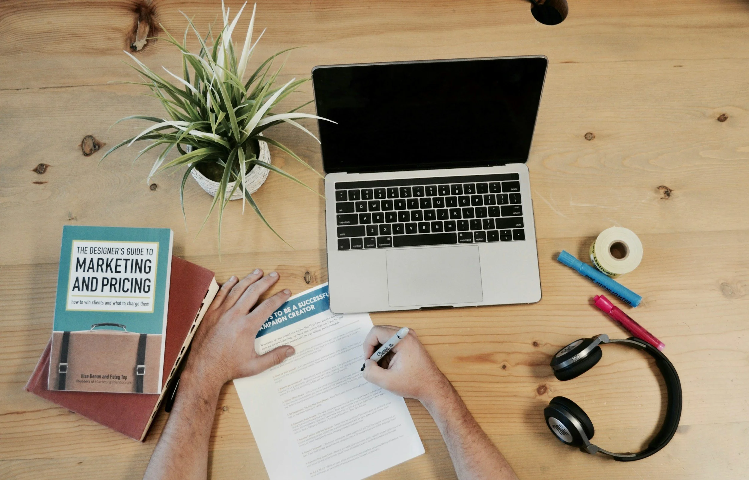 A person working at a wooden desk with a laptop, a potted plant, a marketing book, a printed paper, markers, tape, headphones, and a hand holding a marker writing on the paper.