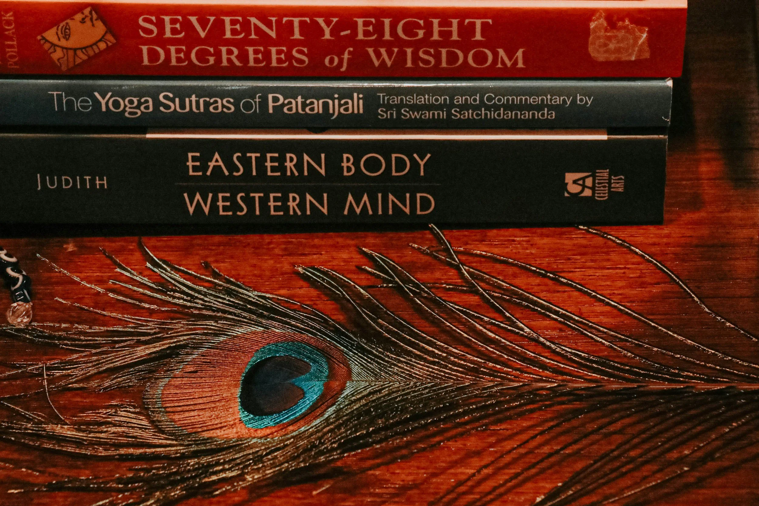 Stack of four books on a wooden surface, with a peacock feather in front. The titles include 'Seventy-Eight Degrees of Wisdom', 'The Yoga Sutras of Patanjali', 'Eastern Body Western Mind', and a partially visible title.