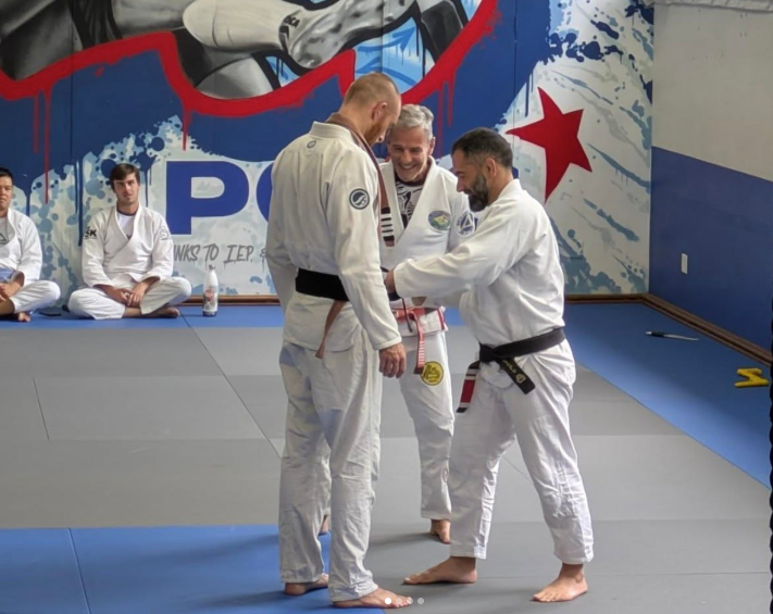 Three men in Brazilian Jiu-Jitsu uniforms standing on a training mat, with one man doing a belt presentation or handshake while the other two observe, in a gym with a graffiti-style wall in the background and two students sitting nearby.