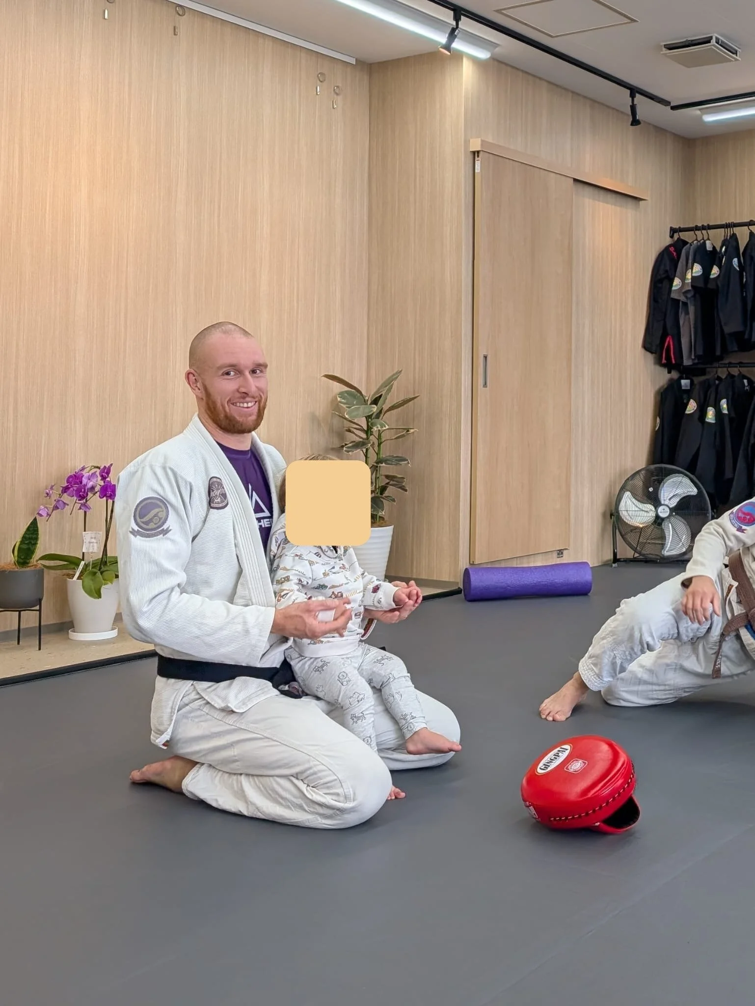 A man in a white martial arts gi kneeling on a black mat, holding a small child, with a woman in a gi seated nearby in a martial arts studio. The studio has wooden walls, a potted plant, and martial arts gear.